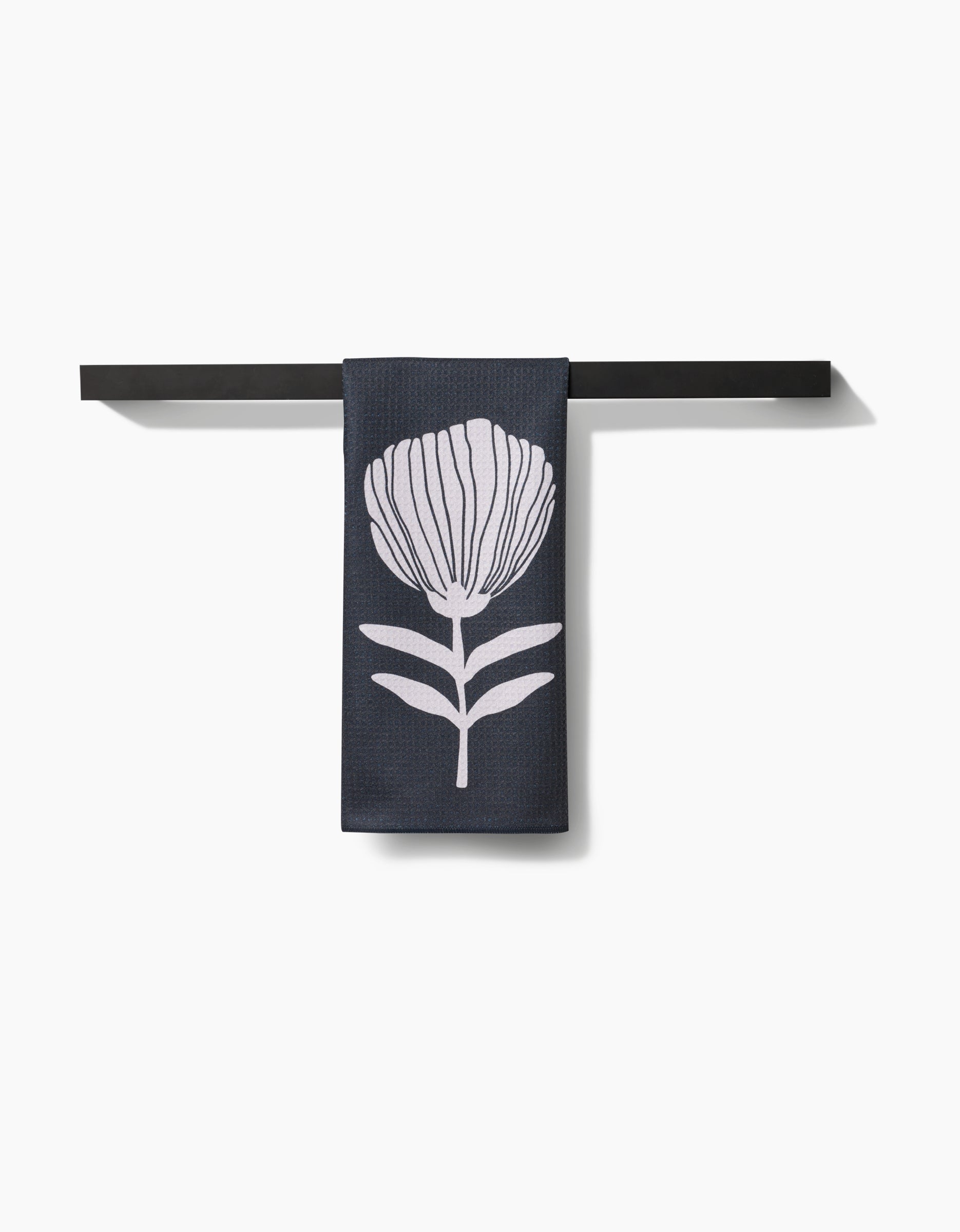The Anna Christian Fika Tea Towel, featuring a white stylized flower on dark Geoweave™, hangs neatly on a simple black bar against a plain white background.