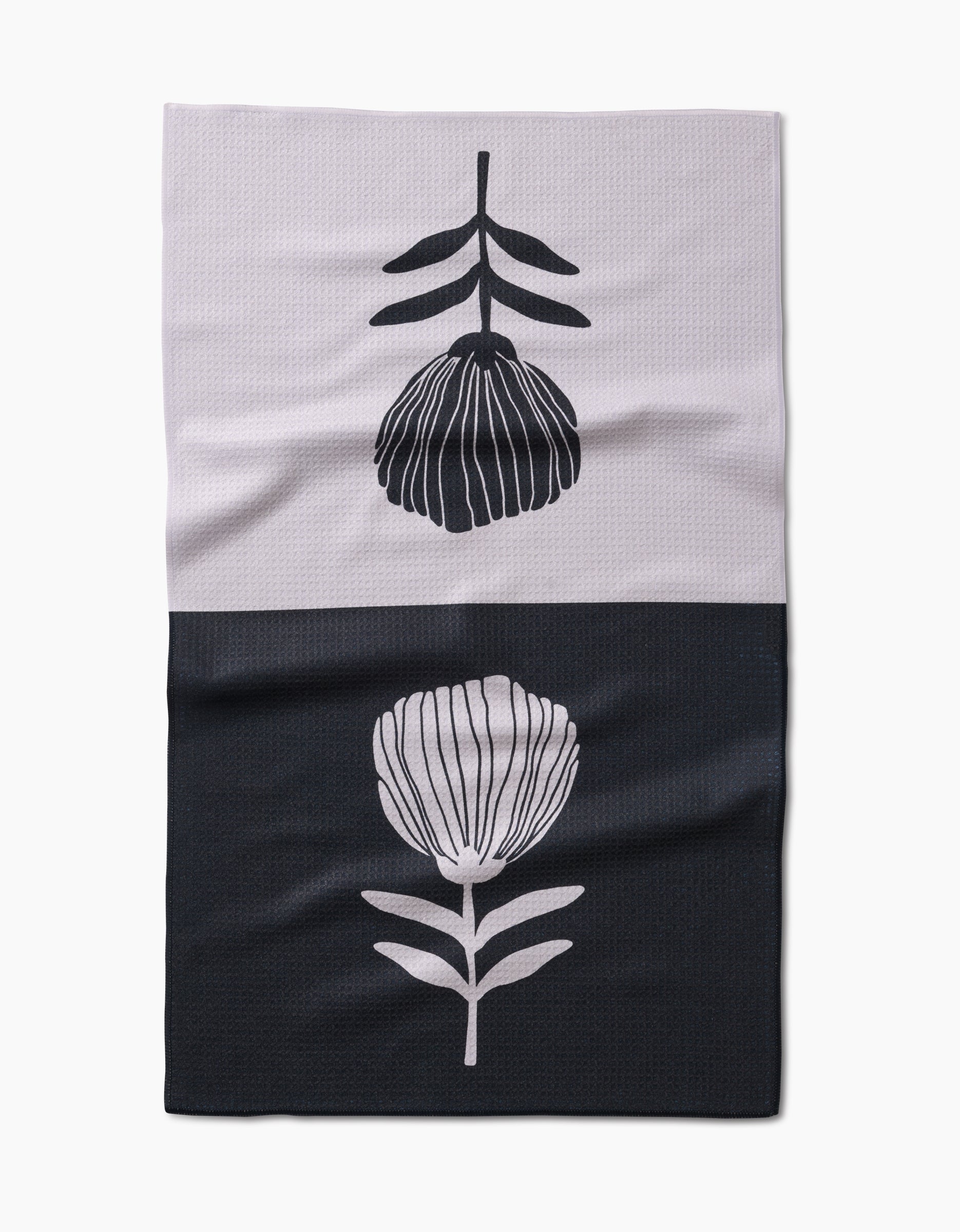 The Fika Tea Towel by Anna Christian features a simple floral design made from recycled materials, with a black flower on light fabric above and a light flower on dark below.