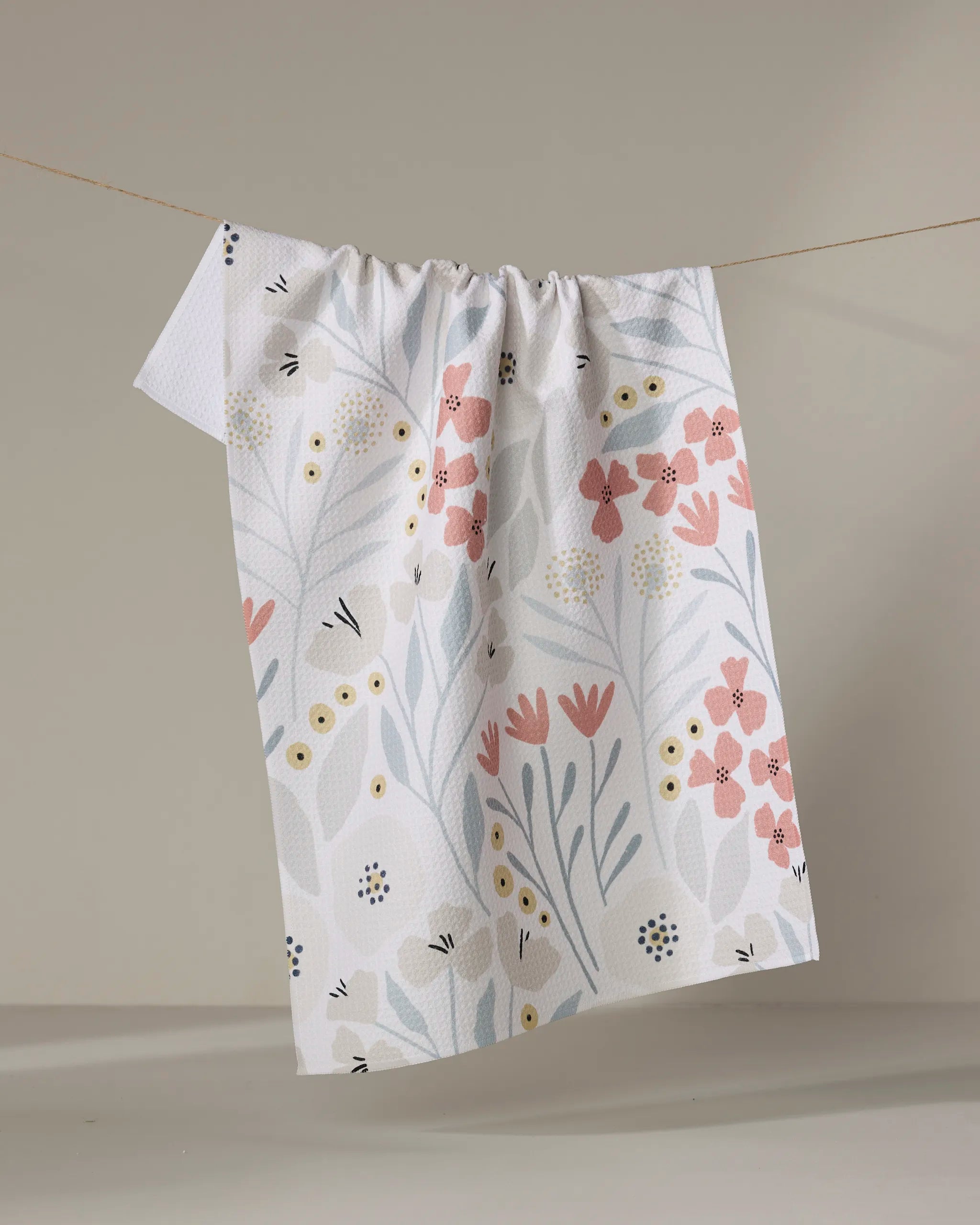 The Fiona Cream tea towel by Amy MacCready, made from recycled materials, displays a pastel floral pattern in pink, yellow, blue, and gray and hangs on a clothesline against a neutral background.