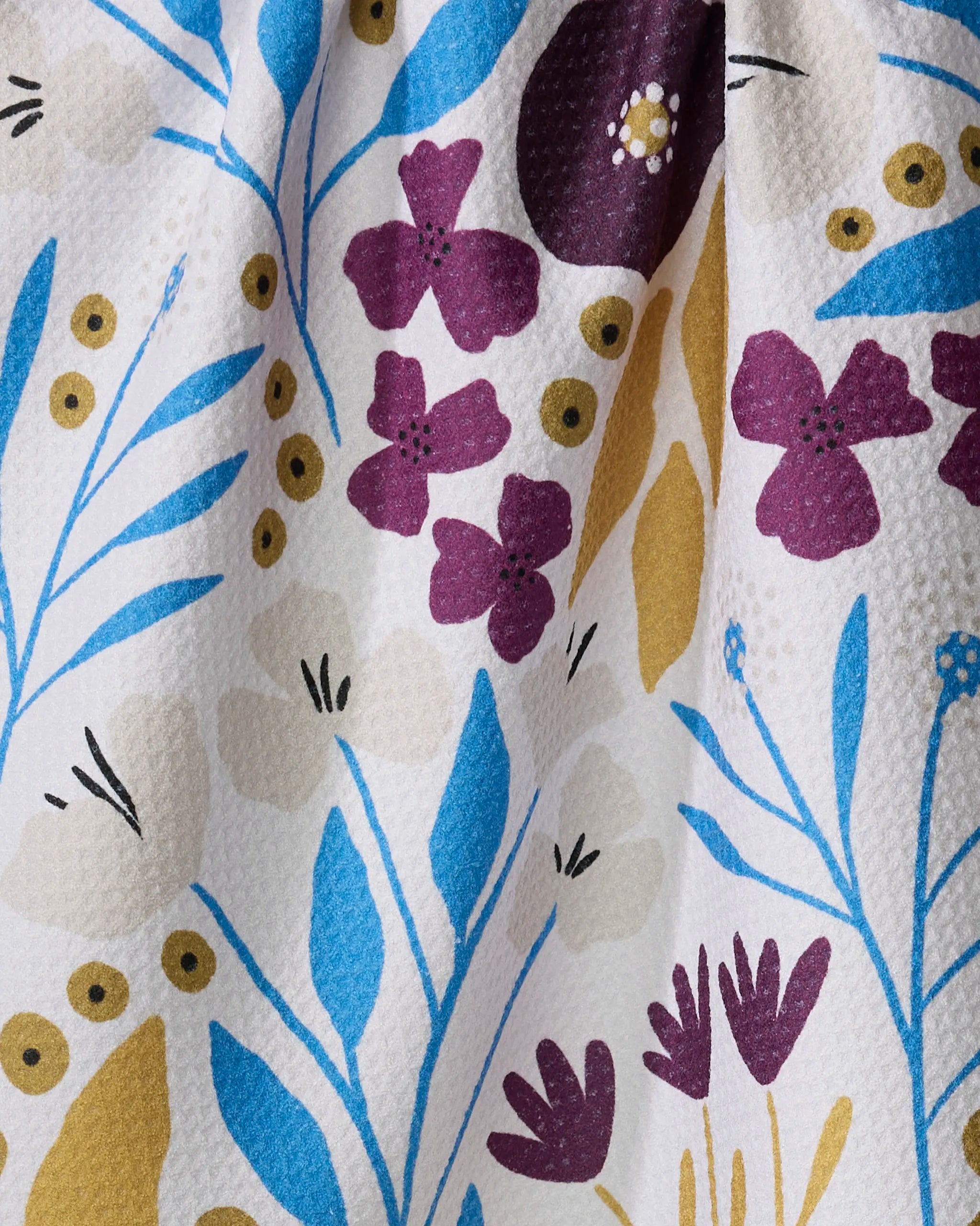 A close-up of the Amy MacCready Fiona Dark Maroon tea towel, showcasing a textured white fabric with a vibrant floral pattern of blue leaves, purple and yellow flowers, small yellow circles, and delicate blue outlines.