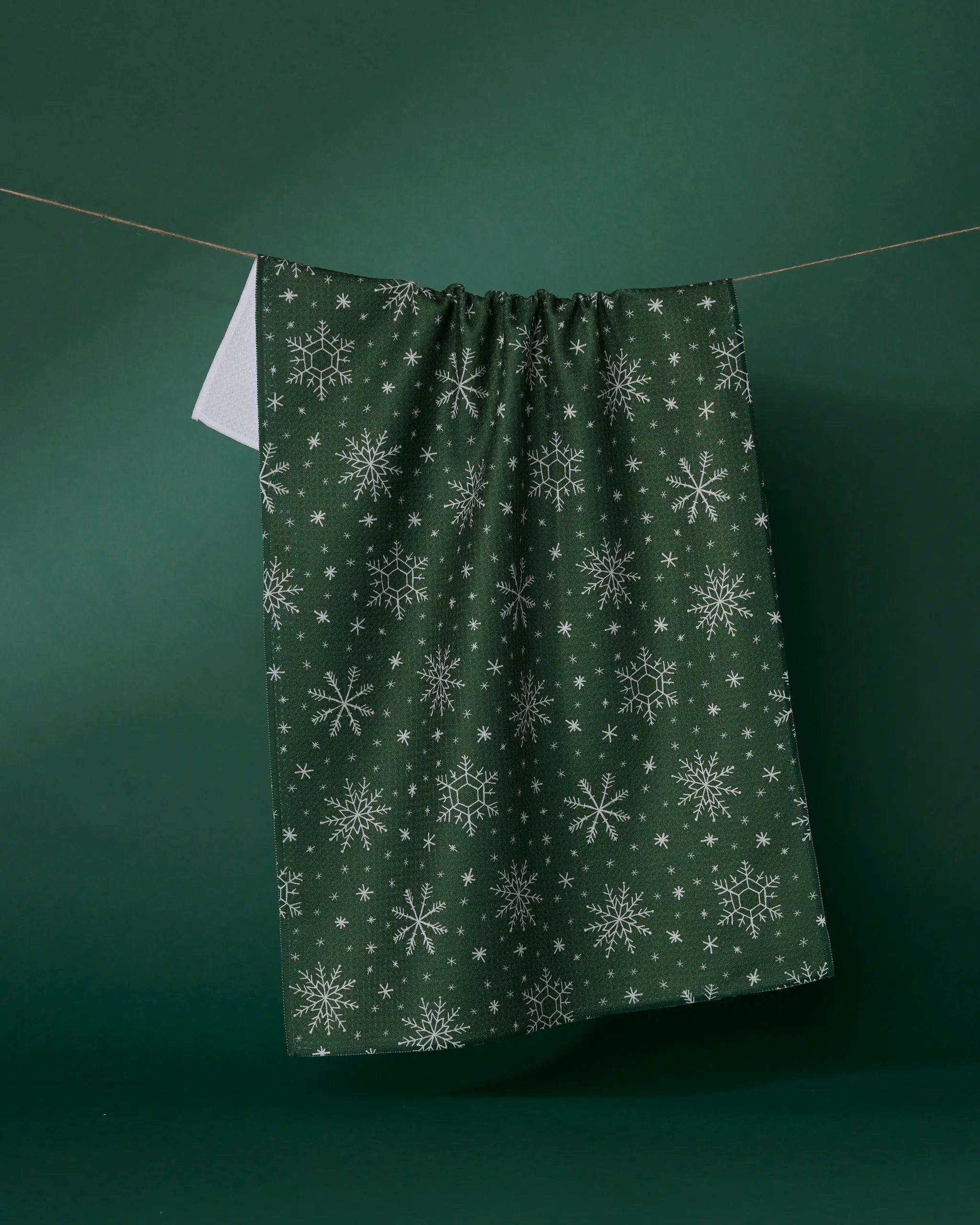 The Geometry Firlight tea towel, an ultra-absorbent dark green towel with white snowflake patterns, hangs on a string against a green background.
