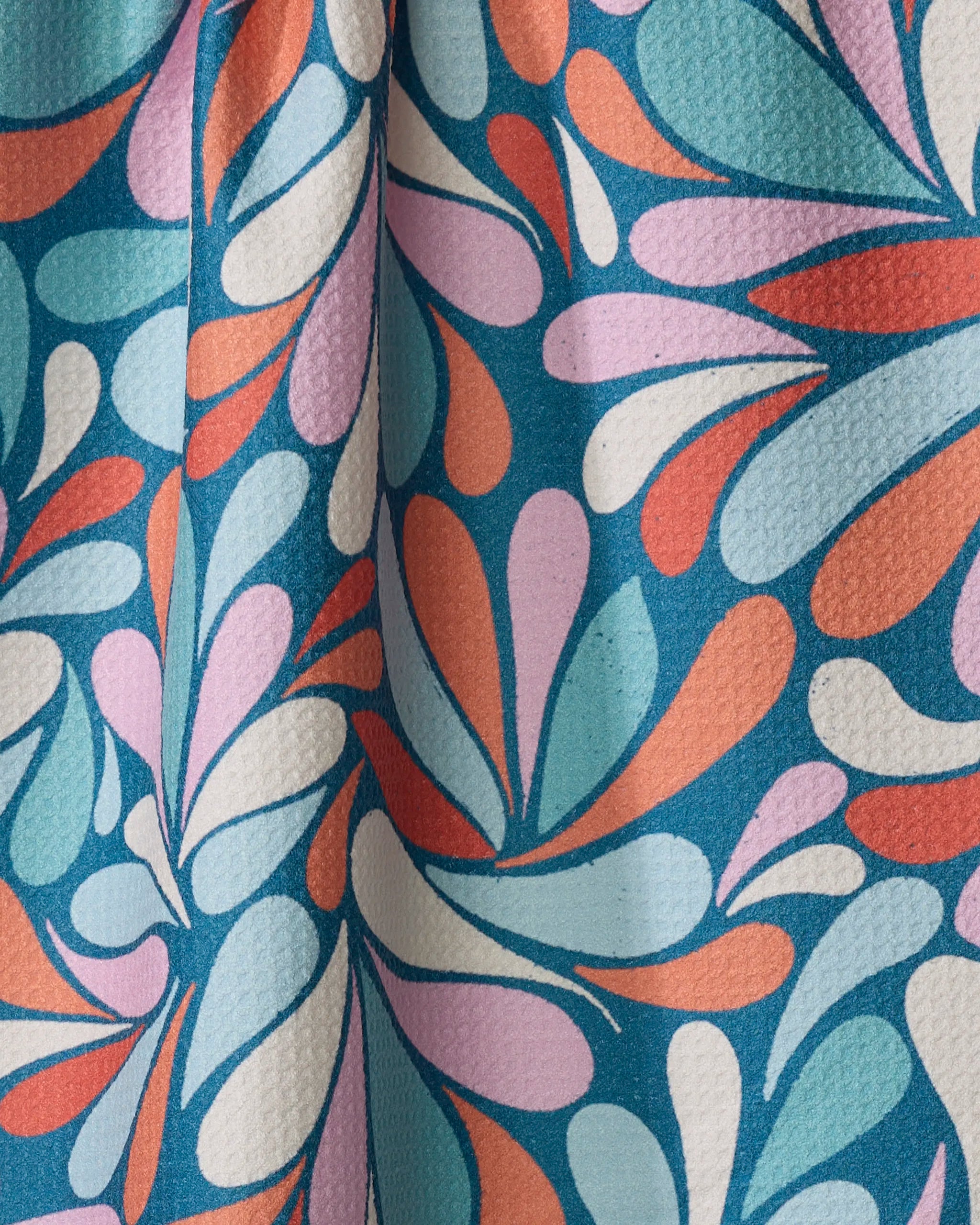 A close-up of Rebecca Woolbright’s First Flight kitchen towel shows its textured fabric and gentle folds, featuring a colorful teardrop pattern in blue, pink, orange, and white on blue, made from recycled materials.