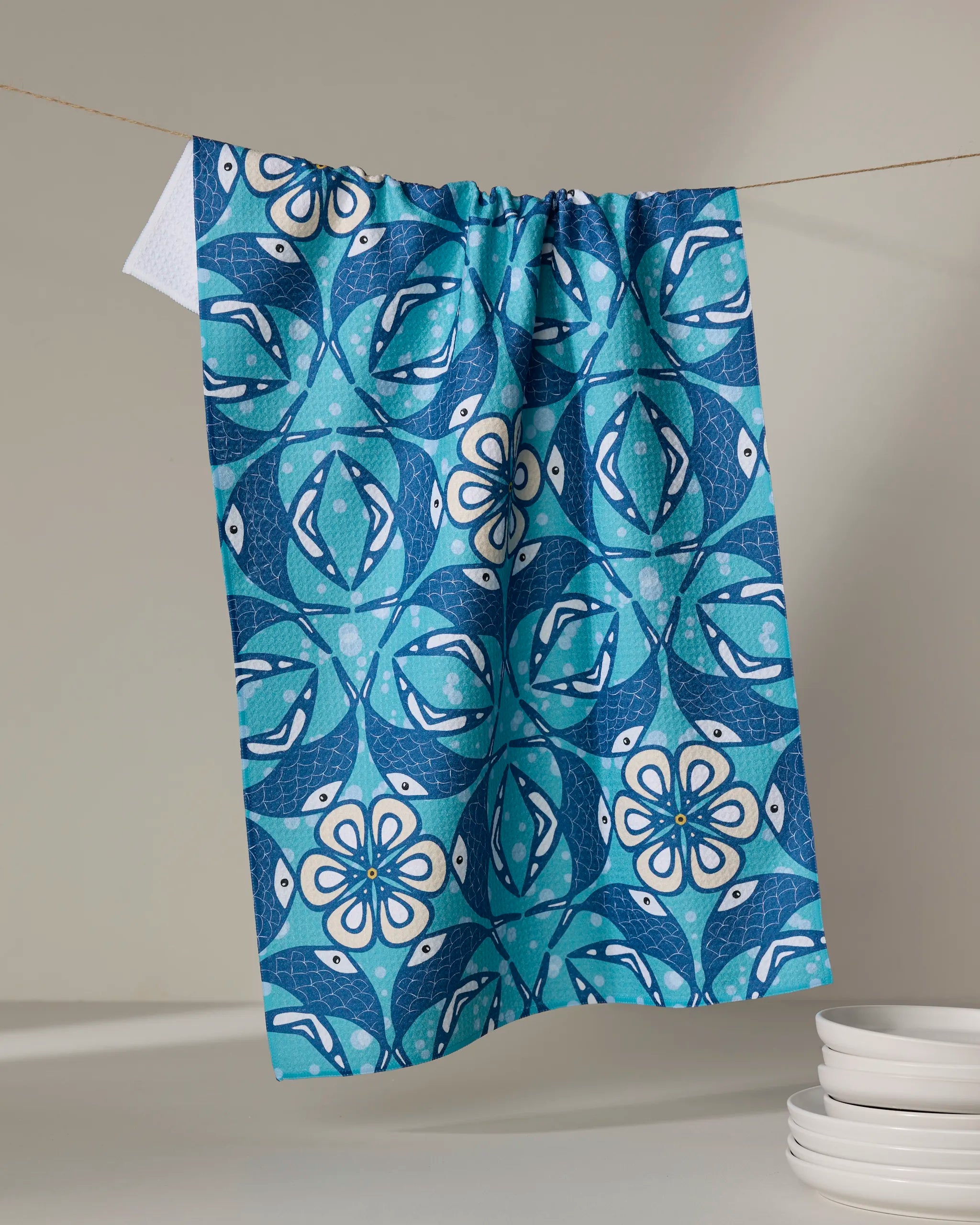The Fish Mandala tea towel by Marta Harvey, featuring a blue and white geometric floral pattern and made from recycled materials, hangs on a string against a neutral background next to a stack of white plates.