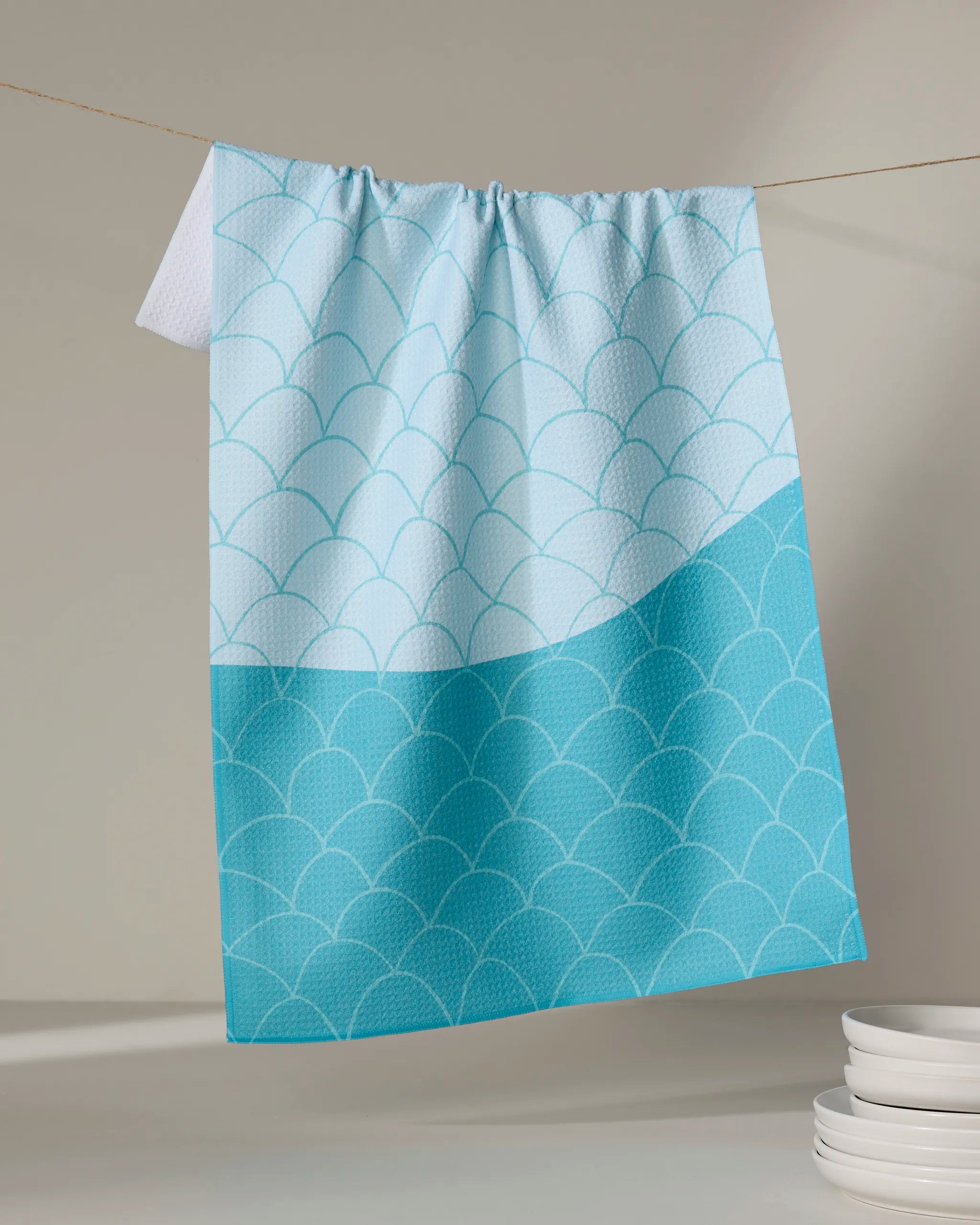 The Marta Harvey Fish Scales tea towel, featuring a light blue and turquoise scallop pattern and made from recycled materials, hangs on a string beside white plates against a neutral background.