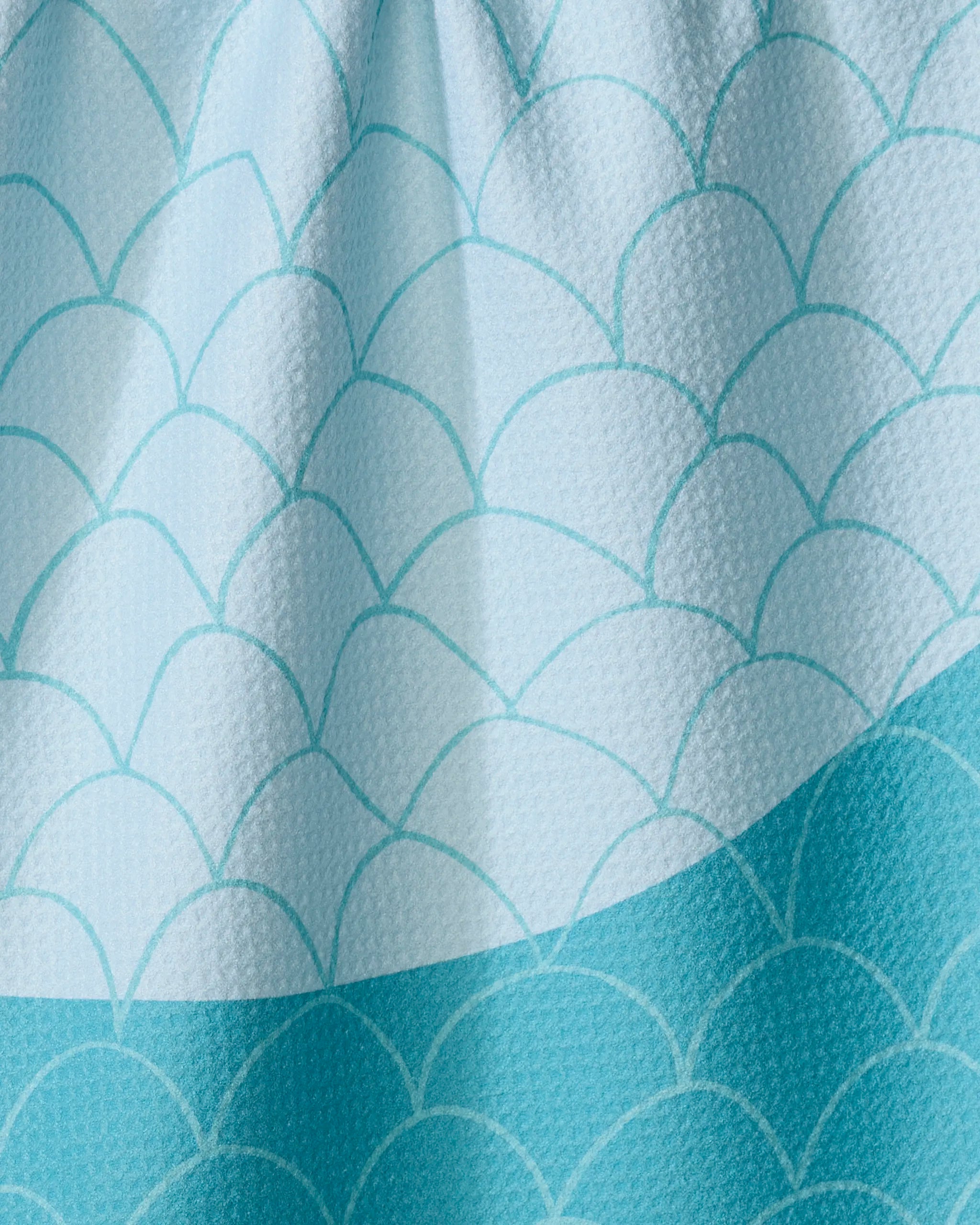 Close-up of the Fish Scales tea towel by Marta Harvey, featuring a textured blue scallop pattern resembling fish scales. Made from recycled materials, this quick-drying towel appears slightly wrinkled at the top.