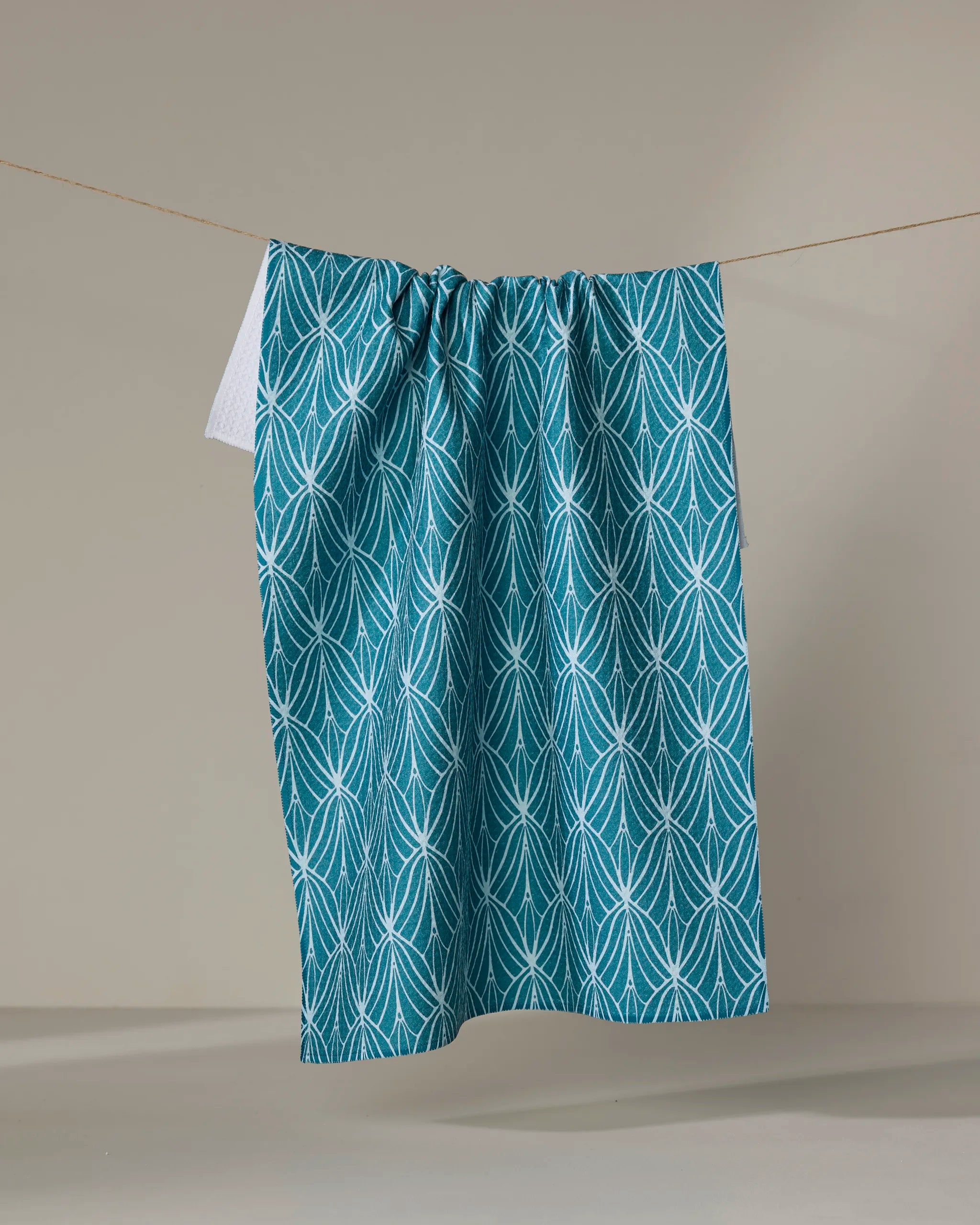 The Marta Harvey Fish Tails tea towel, featuring teal and white fan-like geometric patterns, hangs from a clothesline against a neutral background. Its ultra-absorbent fabric makes it ideal for everyday use.