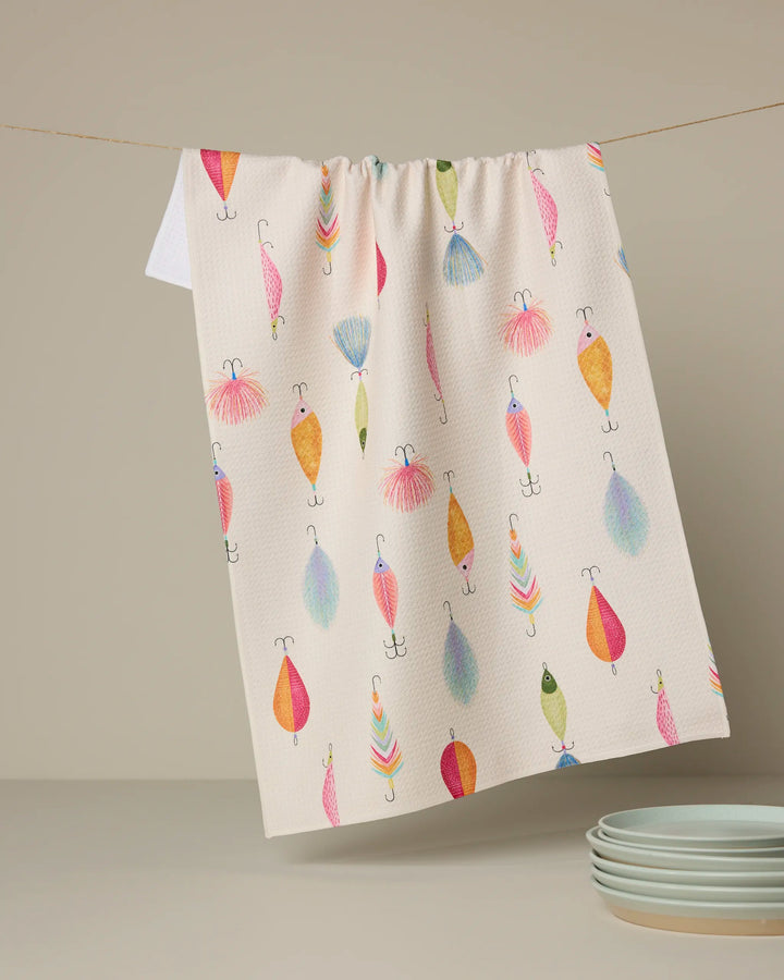 The Geometry "Fishing Lures" kitchen towel, made from recycled materials, features colorful fishing lure illustrations and hangs above four stacked pale green plates against a beige background—perfect for brightening any kitchen.