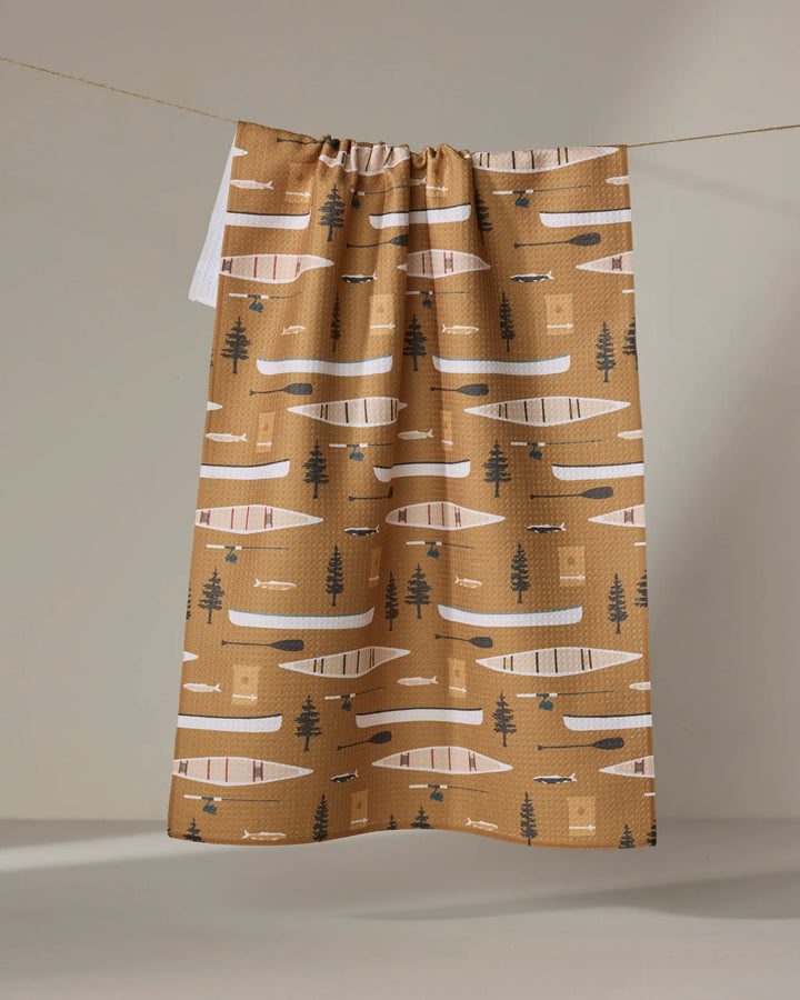 The Coit Creative “Fishing On the Lake” tea towel features an ultra-absorbent brown background with white, black, and beige canoes, paddles, trees, and geometric shapes. It hangs neatly on a line.