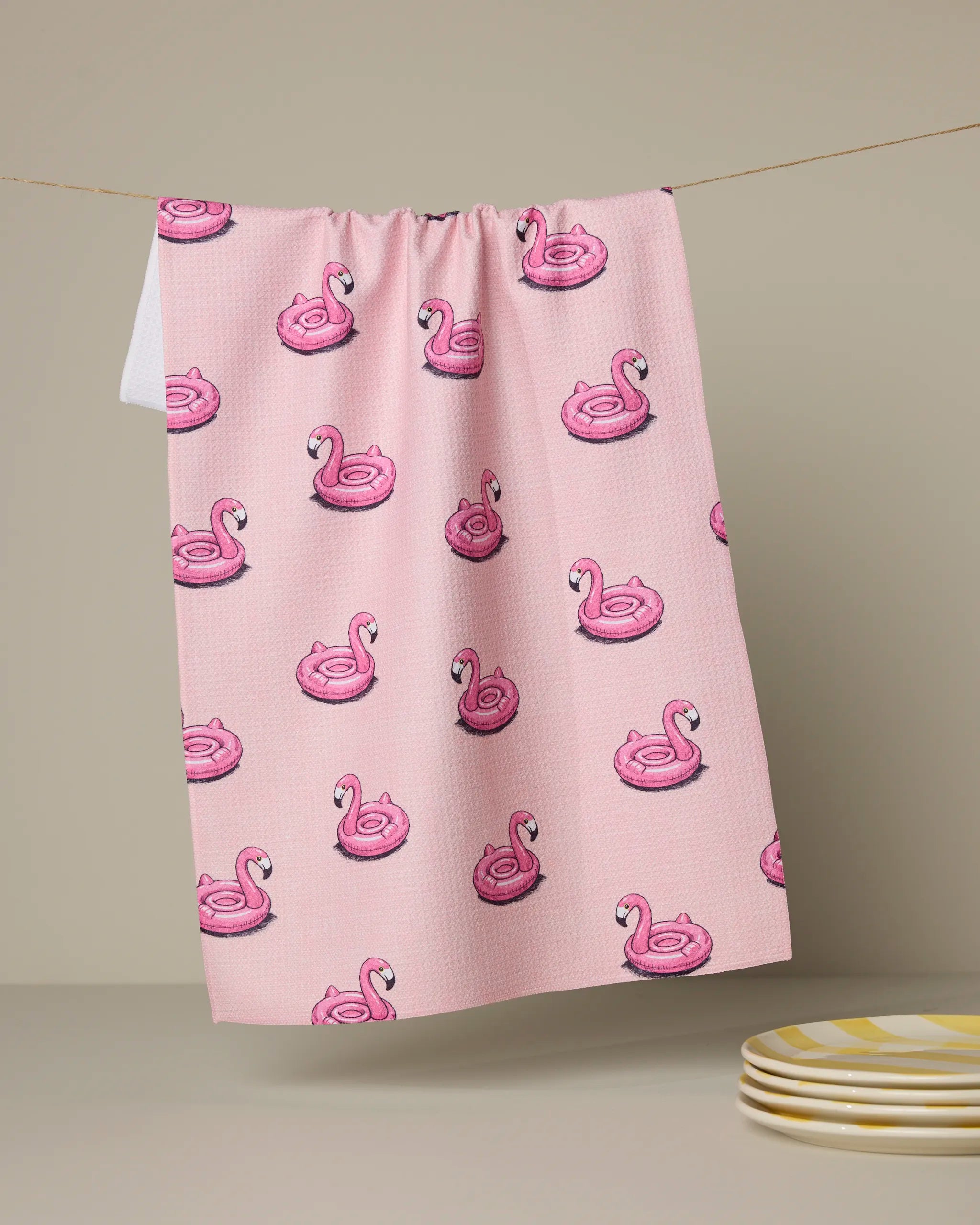 The Geometry Flamingo Floatie kitchen towel, showcasing inflatable pink swan pool floats, hangs above a beige surface with a stack of yellow-striped plates nearby.