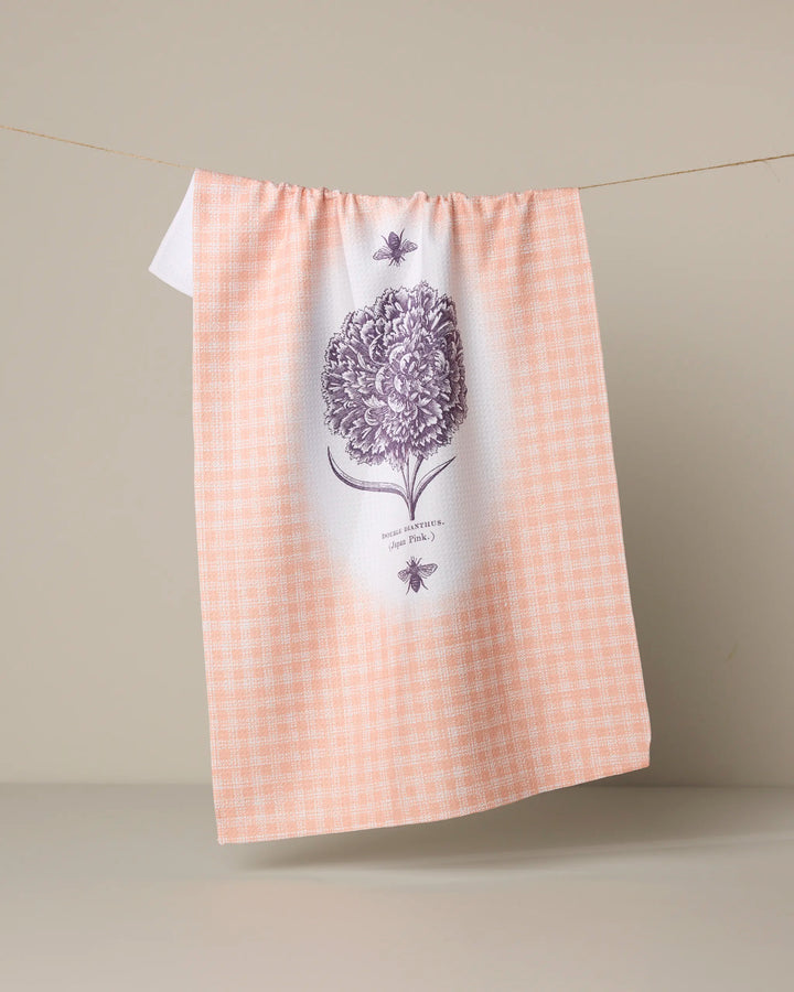 The Geometry "Fleur et Abeille" kitchen tea towel hangs on a line. Made from recycled materials, it features a peach and white checkered pattern, a purple flower with two bees, and text below, set against a neutral beige background.