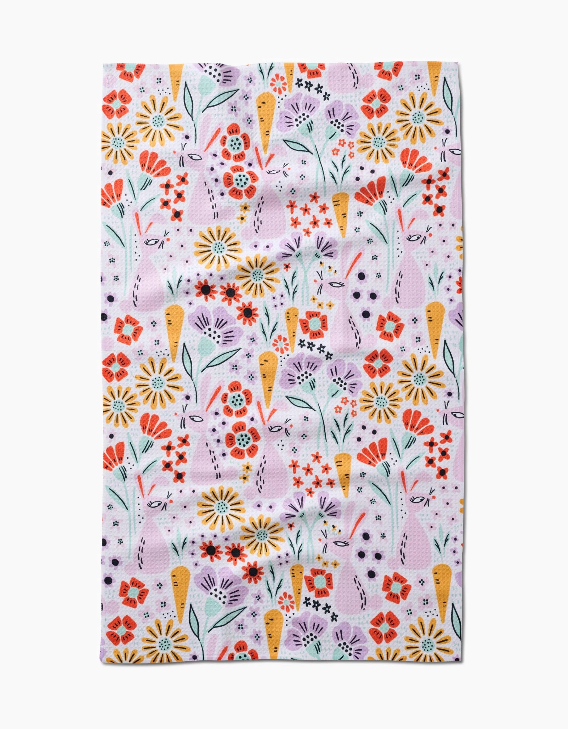 The Floppsie tea towel by Amy MacCready features a quick-drying fabric with a whimsical, repetitive floral pattern of red, yellow, and purple flowers with green stems and leaves on a light pink background.