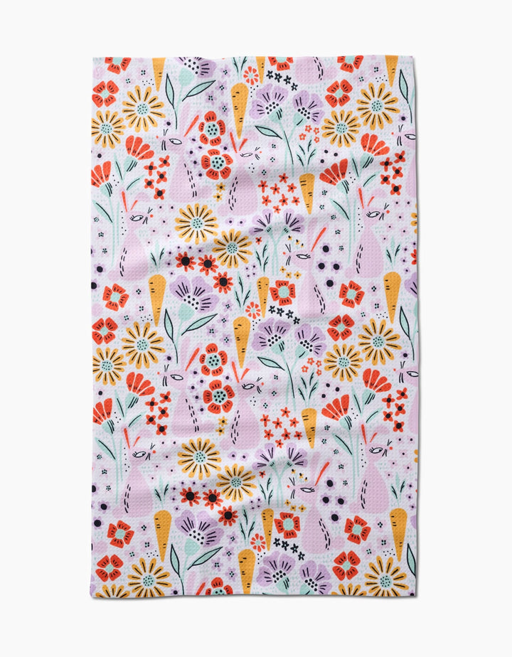 The Floppsie tea towel by Amy MacCready features a quick-drying fabric with a whimsical, repetitive floral pattern of red, yellow, and purple flowers with green stems and leaves on a light pink background.