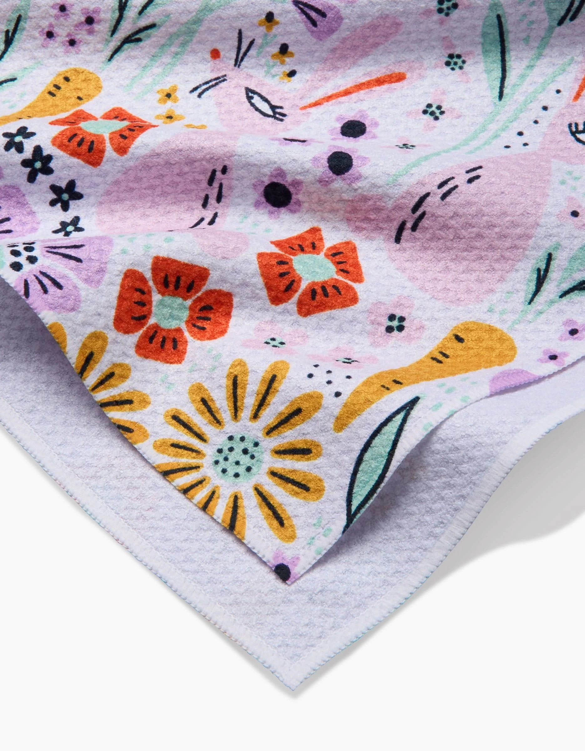 A close-up of the Floppsie tea towel by Amy MacCready, made from recycled materials, featuring a colorful floral print with red, yellow, and purple flowers on a textured lavender background. The edge appears in the bottom left corner.