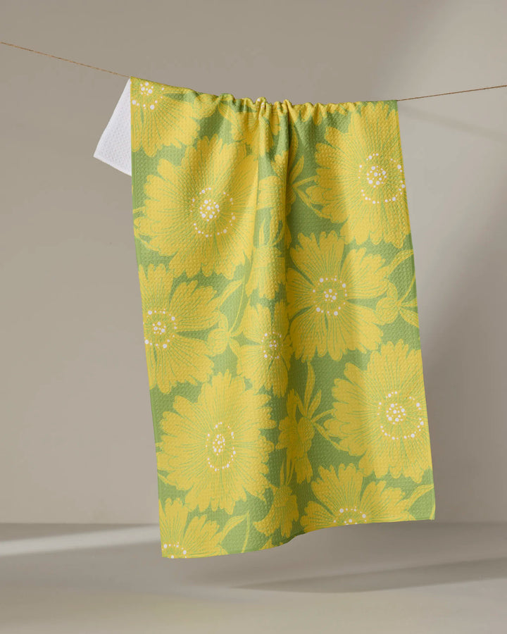 The Dabney Lee Flora towel, featuring a bold sunflower pattern in yellow and green, hangs on a clothesline with a hint of white fabric visible behind it.