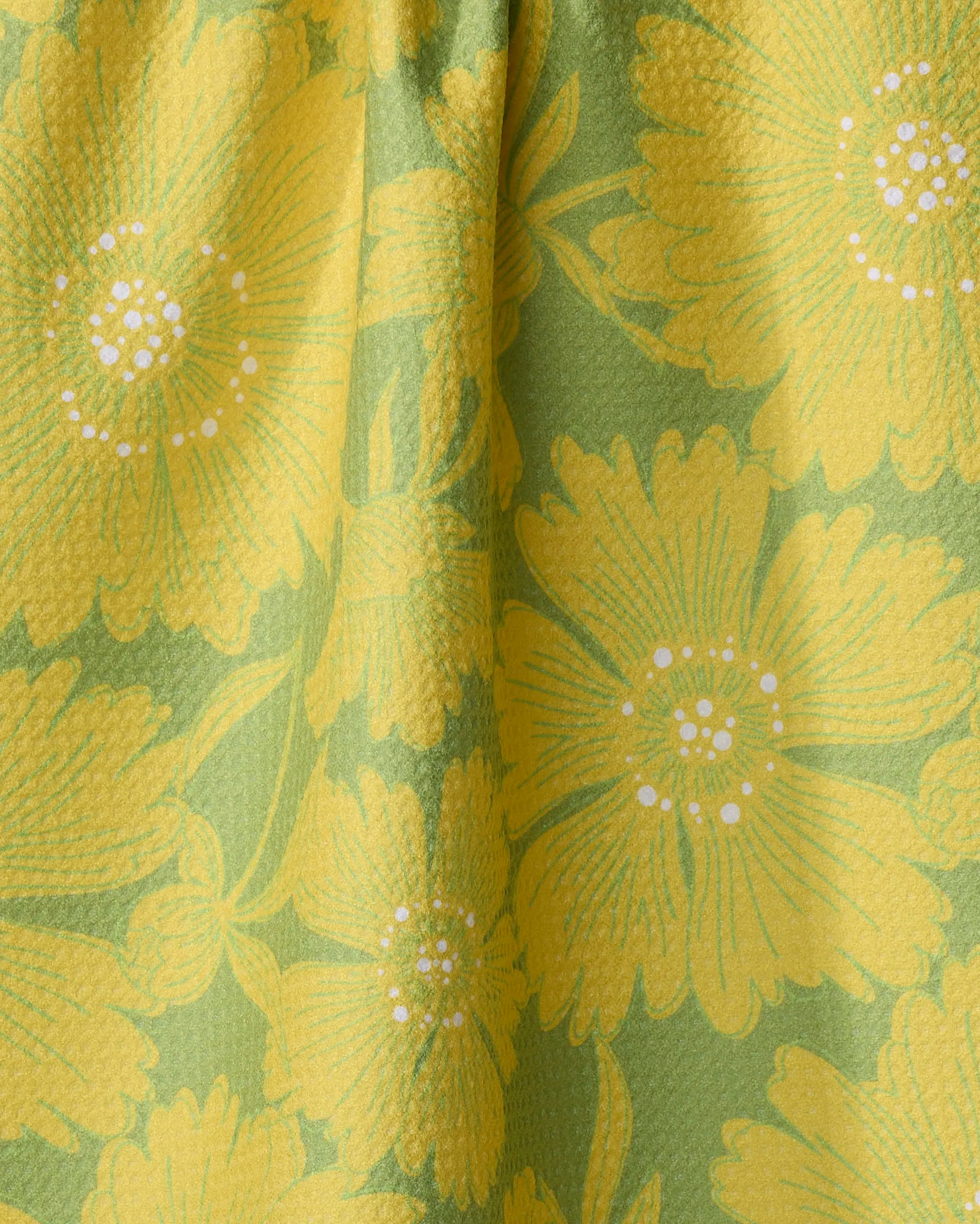 The Dabney Lee Flora kitchen towel features a vibrant, summery floral pattern of yellow flowers with white centers on green textured recycled fabric.