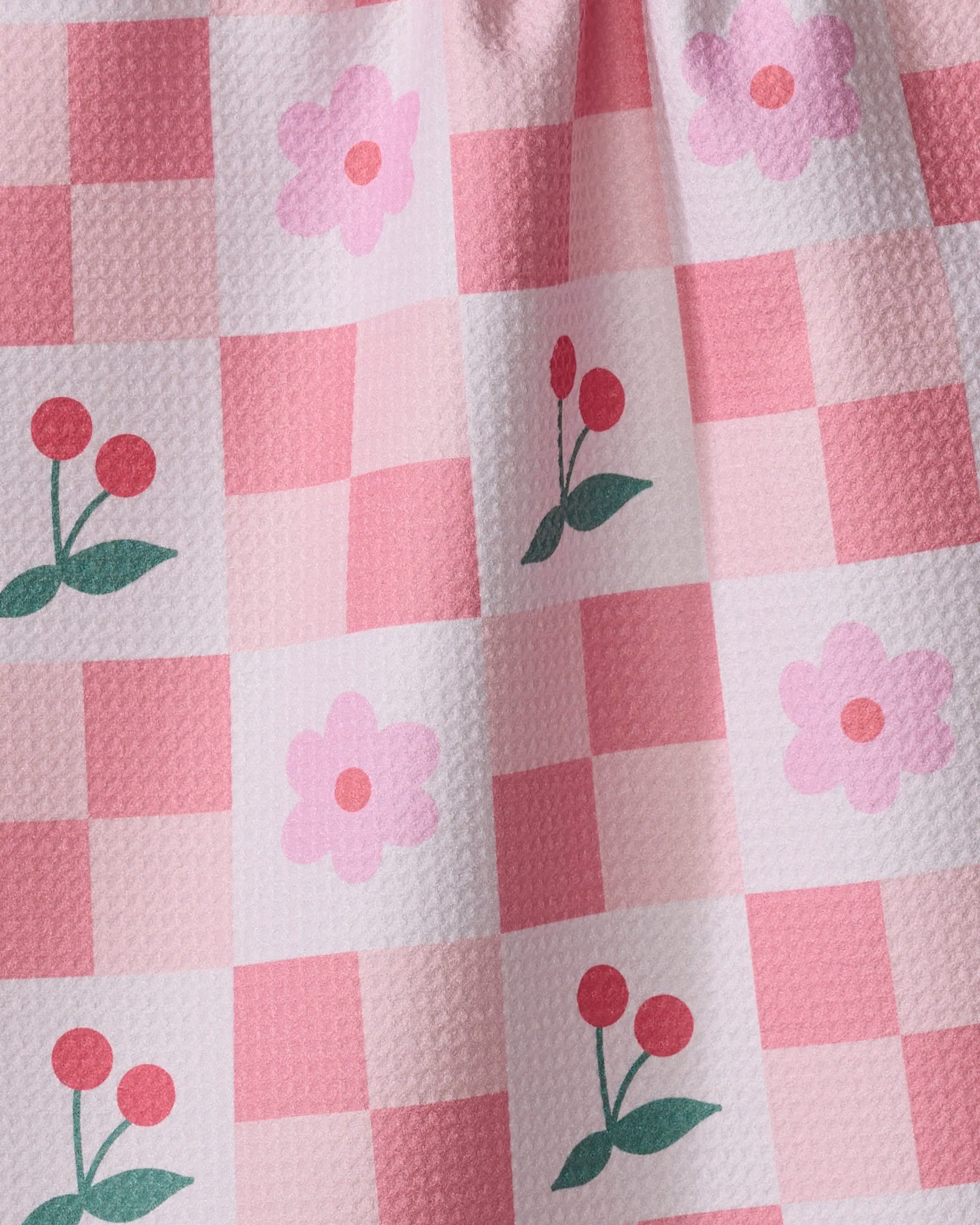 The "Floral and Cherries" tea towel by Letters By Shells features a pink and white checkered design, textured surface, and charming illustrations of pink flowers and red cherries with green leaves. Made from recycled materials.