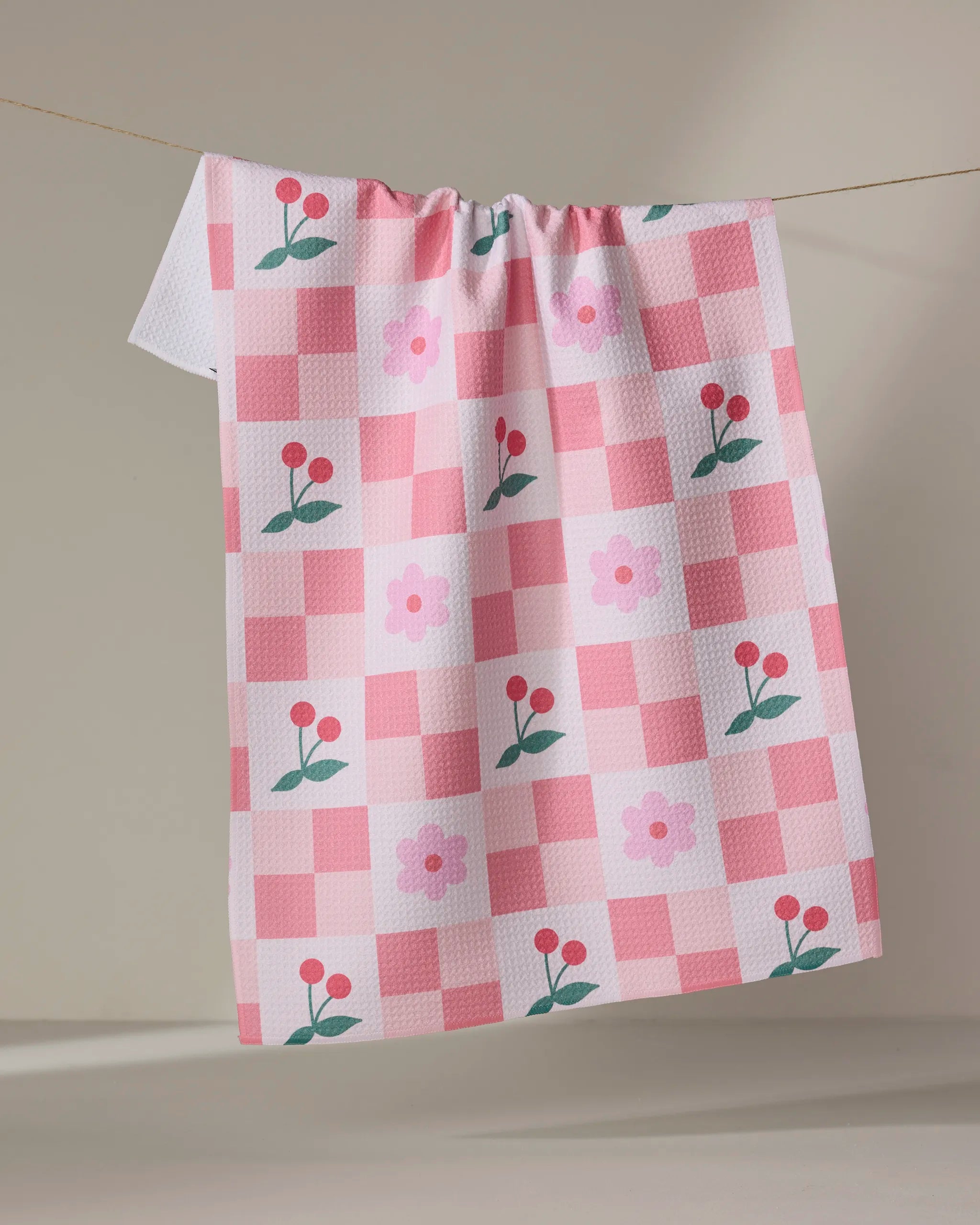Floral and Cherries tea towel by Letters By Shells, featuring pink and white checks with flower and berry designs, crafted from recycled materials, hangs on a clothesline against a simple light background.