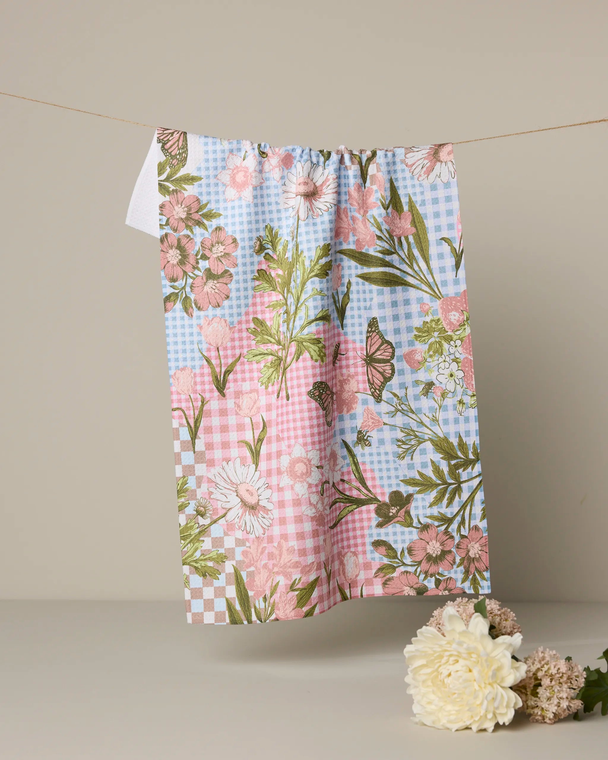 The Geometry Floral Collage tea towel, featuring pink, white, and green flowers with butterflies and blue-pink checks, hangs on a string; cream-colored flowers lie below.