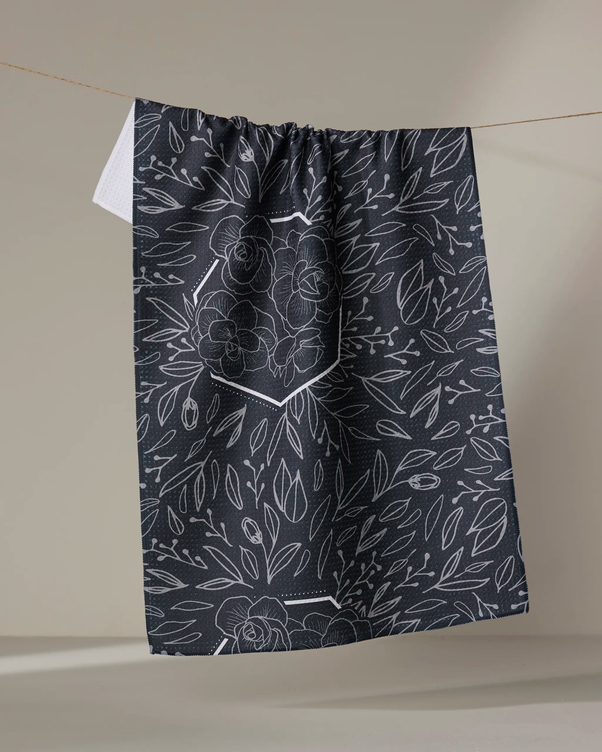 The Rosa Kichline "Floral Explosion" tea towel, featuring a white floral and leafy pattern on a dark background, hangs on a line against a plain, neutral backdrop.