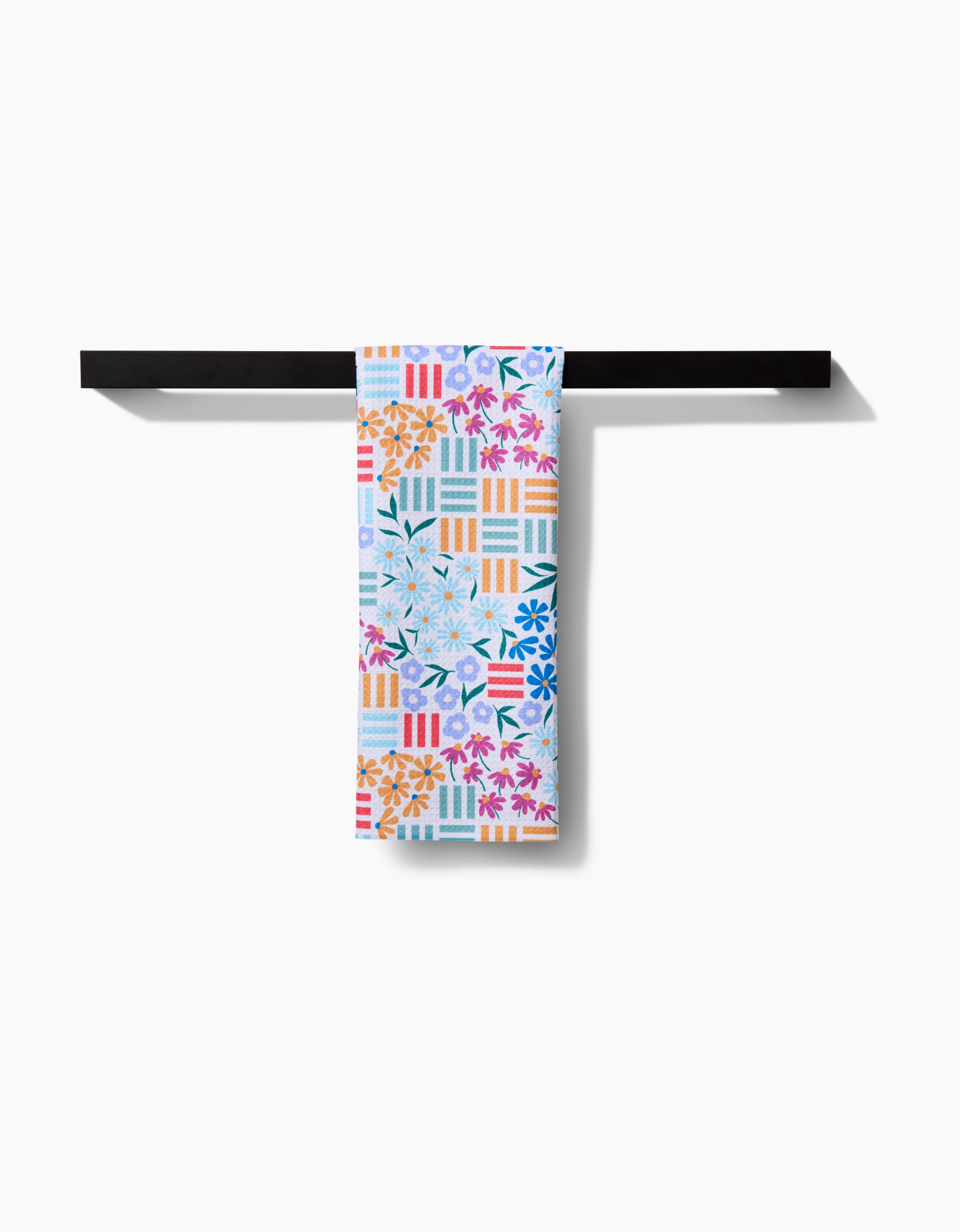 The Floral Garden tea towel by Charlie Rowan Designs, featuring a geometric and floral Geoweave pattern, hangs neatly on a black towel bar against a white background.