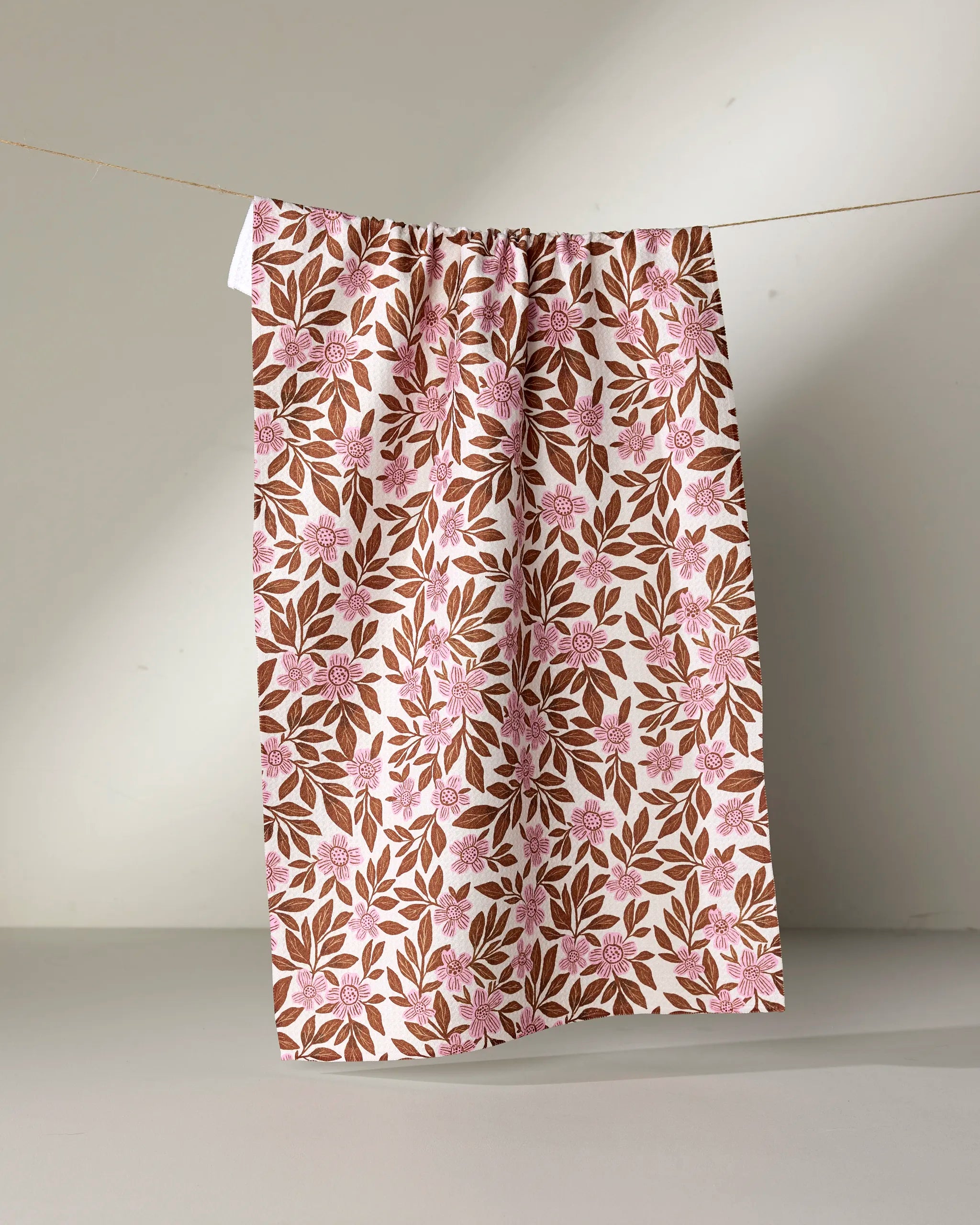 The Lisa Nelson "Floral Meadow" tea towel, featuring pink flowers and brown leaves on ultra-absorbent white fabric, hangs from a clothesline against a neutral background.
