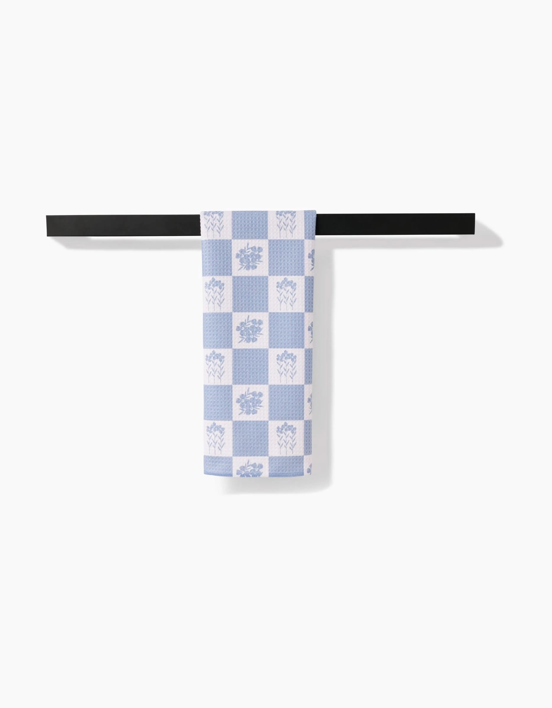 The Floral Patchwork tea towel by Charlie Rowan Designs, featuring light blue and white checkered florals on Geoweave fabric, hangs neatly on a black bar against a plain white background.