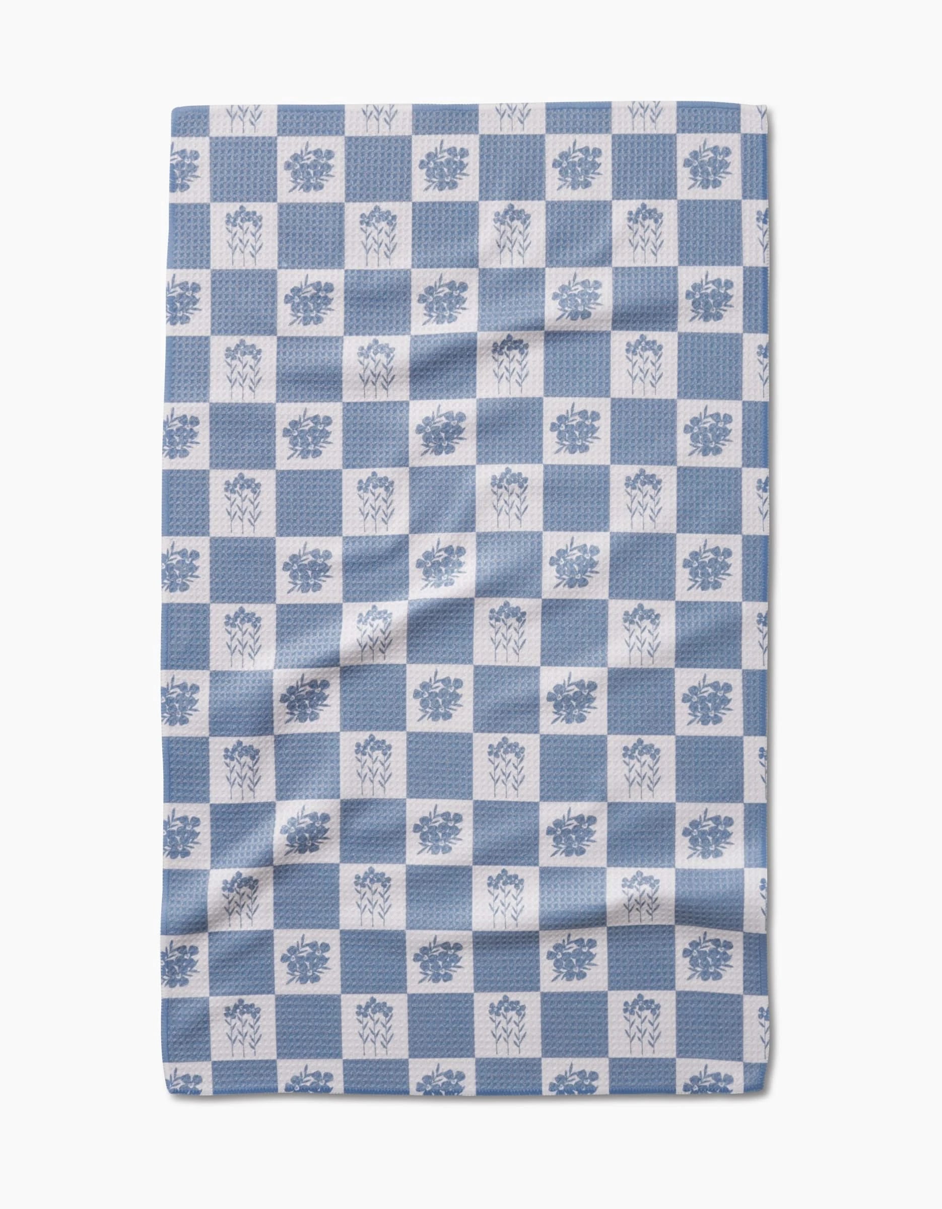The Floral Patchwork tea towel by Charlie Rowan Designs features a blue and white checkered pattern with alternating floral squares. Made from recycled materials, its slightly wrinkled fabric stands out against a white background.