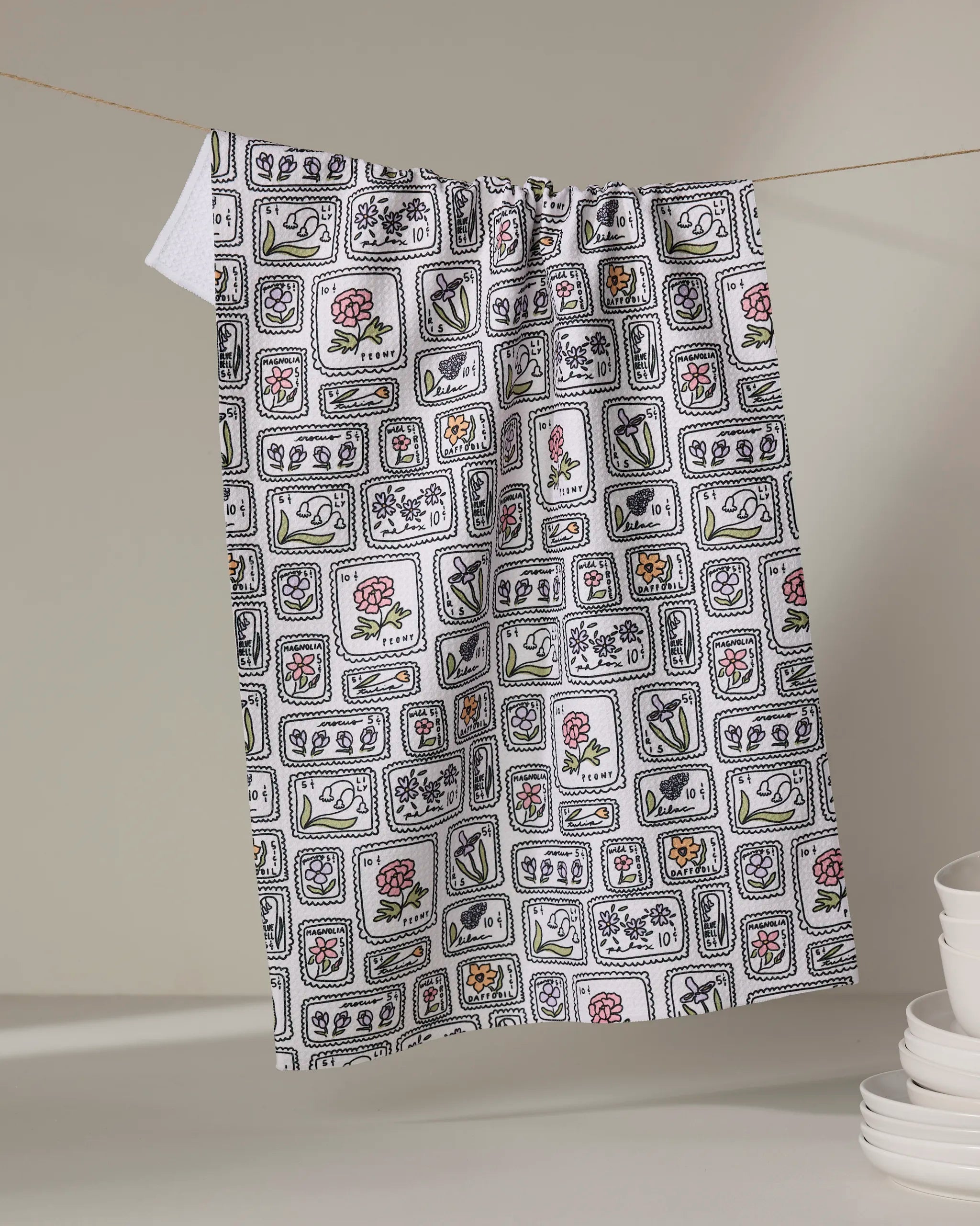 The Kayla Ann Floral Postage Stamps tea towel features a white base with a black grid and colorful floral illustrations. Made from recycled materials, it hangs on a string against a neutral background near stacked white dishes.