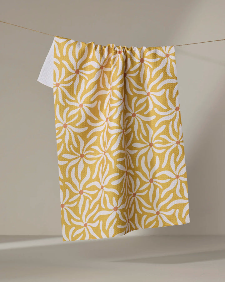 The Gerdadzy "Floral Spell" tea towel, featuring a yellow and white floral pattern and made from recycled materials, hangs on a clothesline against a plain neutral background.
