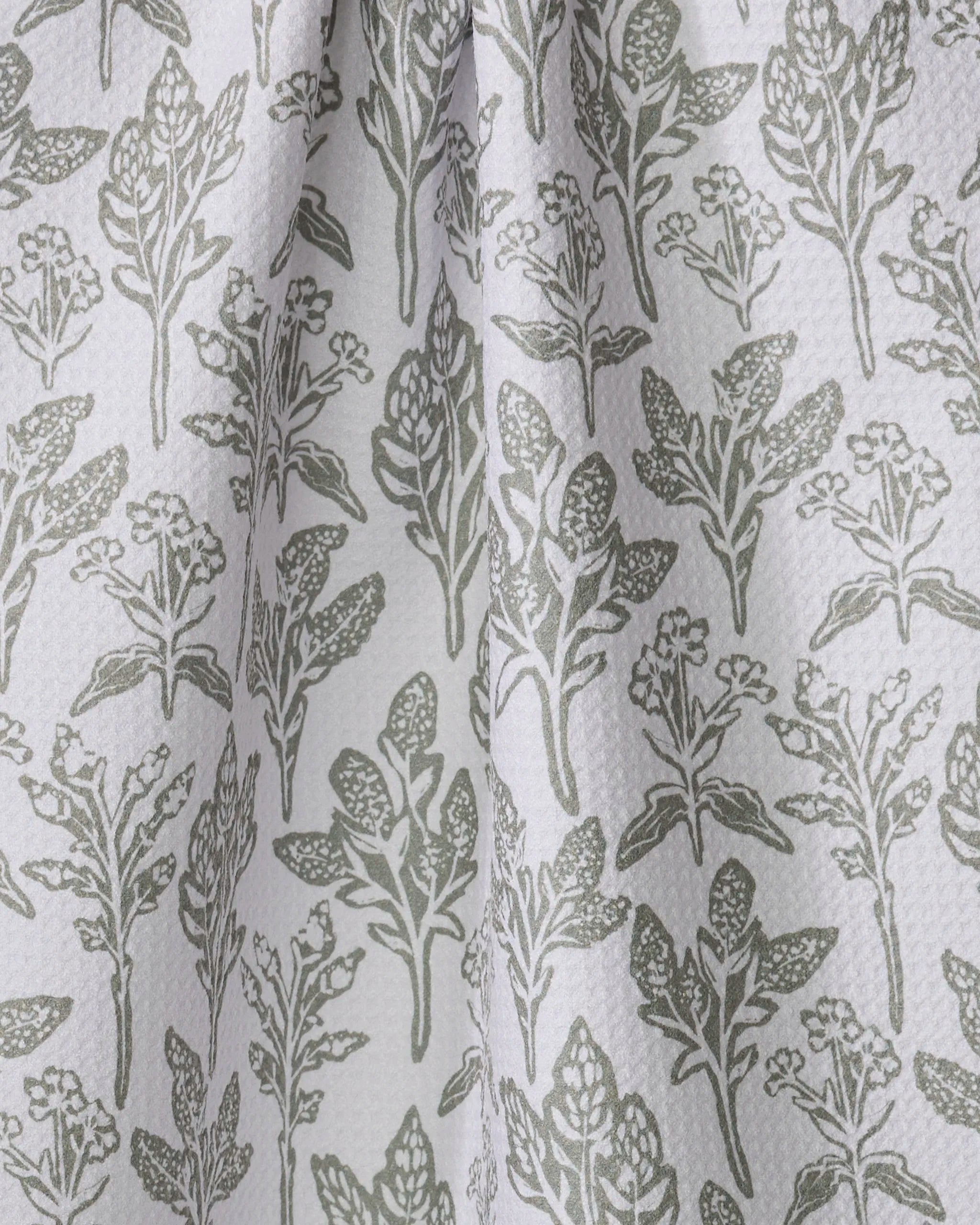 The “Floral Stamp” kitchen towel by Hufton Studio features a white textured fabric with a repeating pattern of green botanical leaves, stems, and small flowers, plus gentle vertical folds.