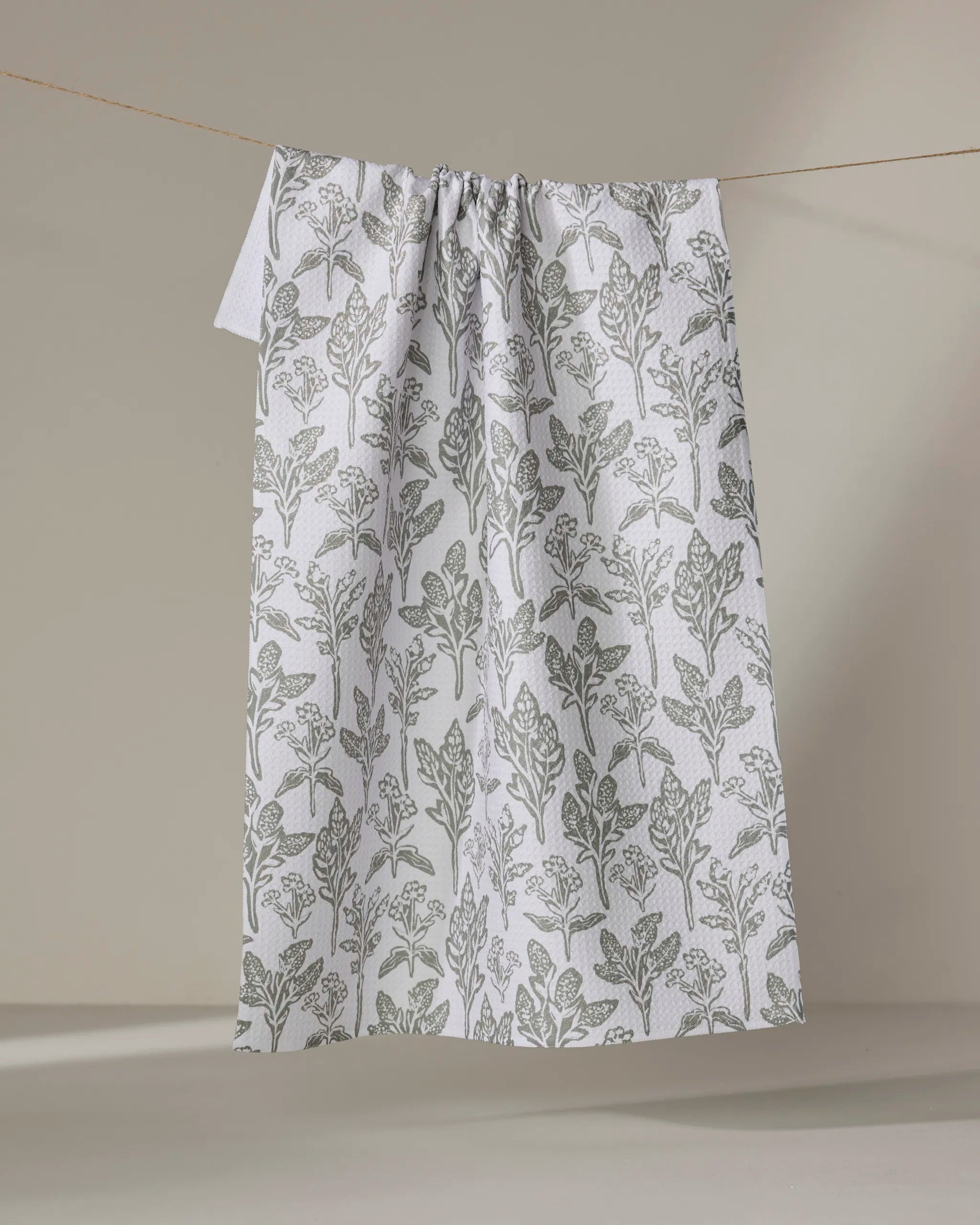 The Hufton Studio "Floral Stamp" kitchen towel, crafted from recycled materials and featuring a white background with a gray botanical leaf pattern, hangs on a string against a neutral beige backdrop.