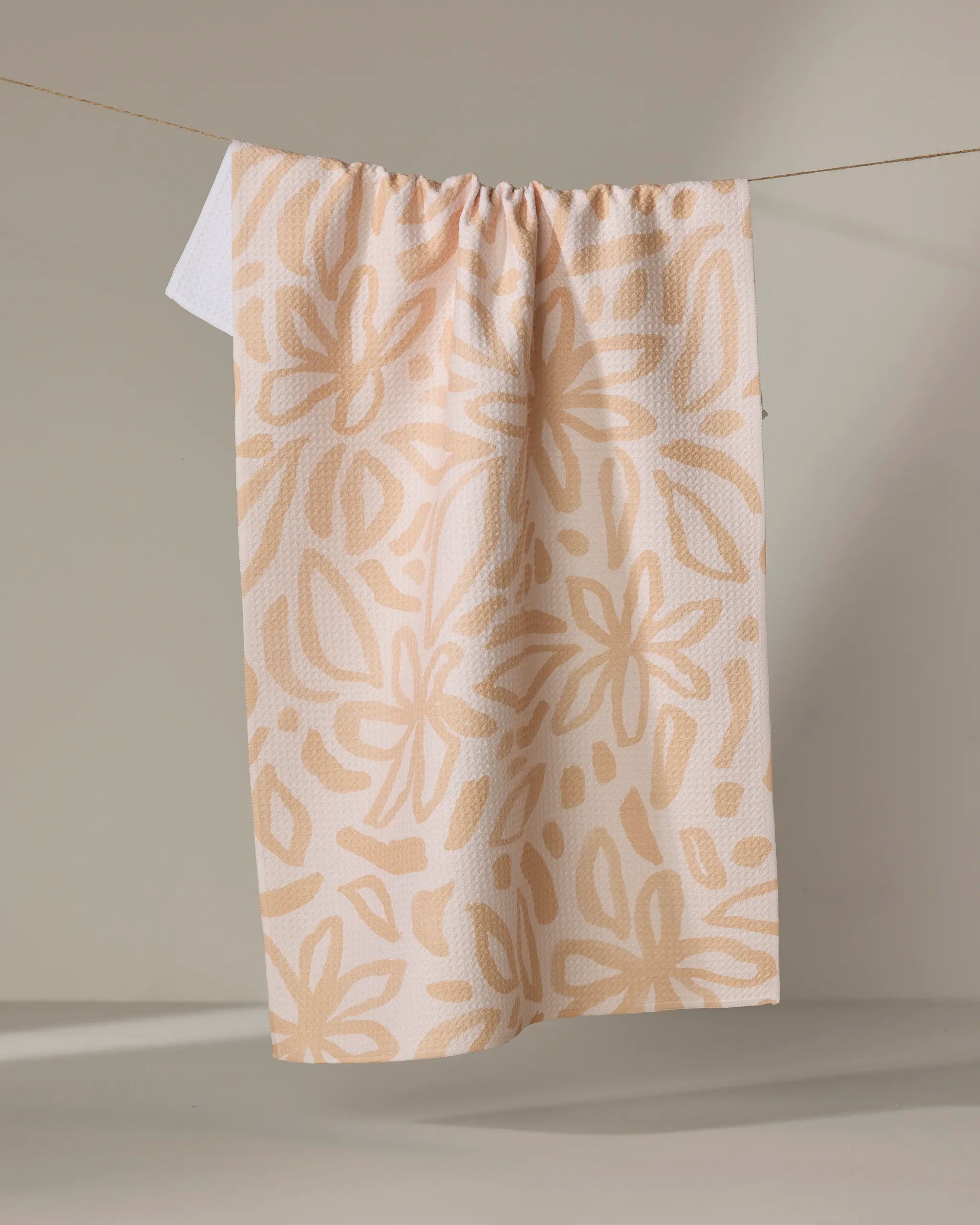 The Jen B. Peters Floral towel, an eco-friendly beige kitchen towel with a large abstract light tan floral pattern, hangs from a string against a neutral background.