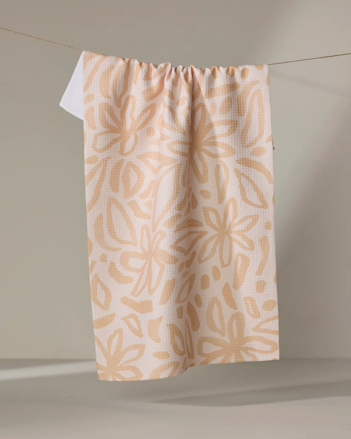 The Jen B. Peters Floral towel, an eco-friendly beige kitchen towel with a large abstract light tan floral pattern, hangs from a string against a neutral background.