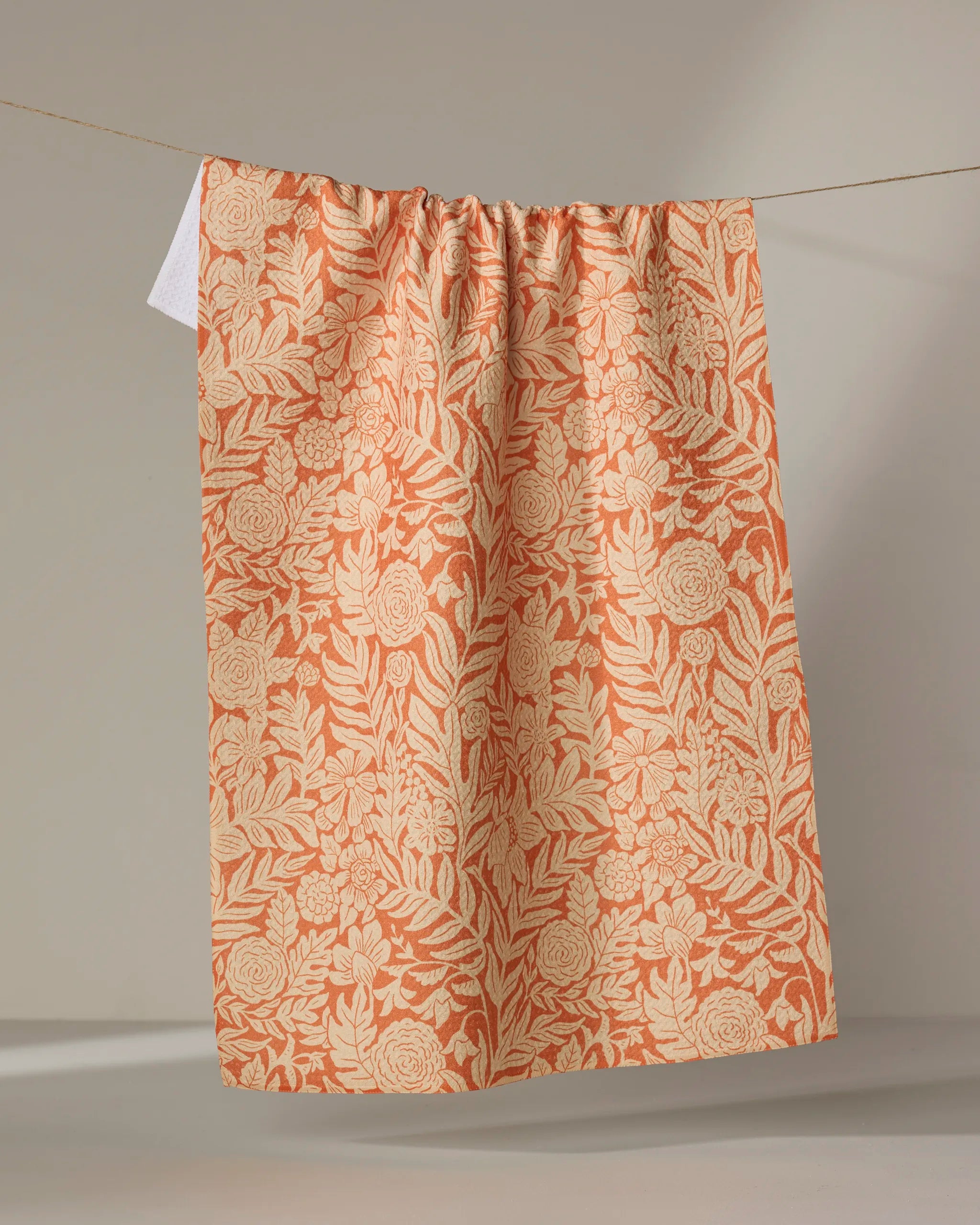 The Florence kitchen towel by Katie O'Shea, in peach with a cream floral and leafy pattern, is made from recycled materials and hangs from a string against a neutral background.