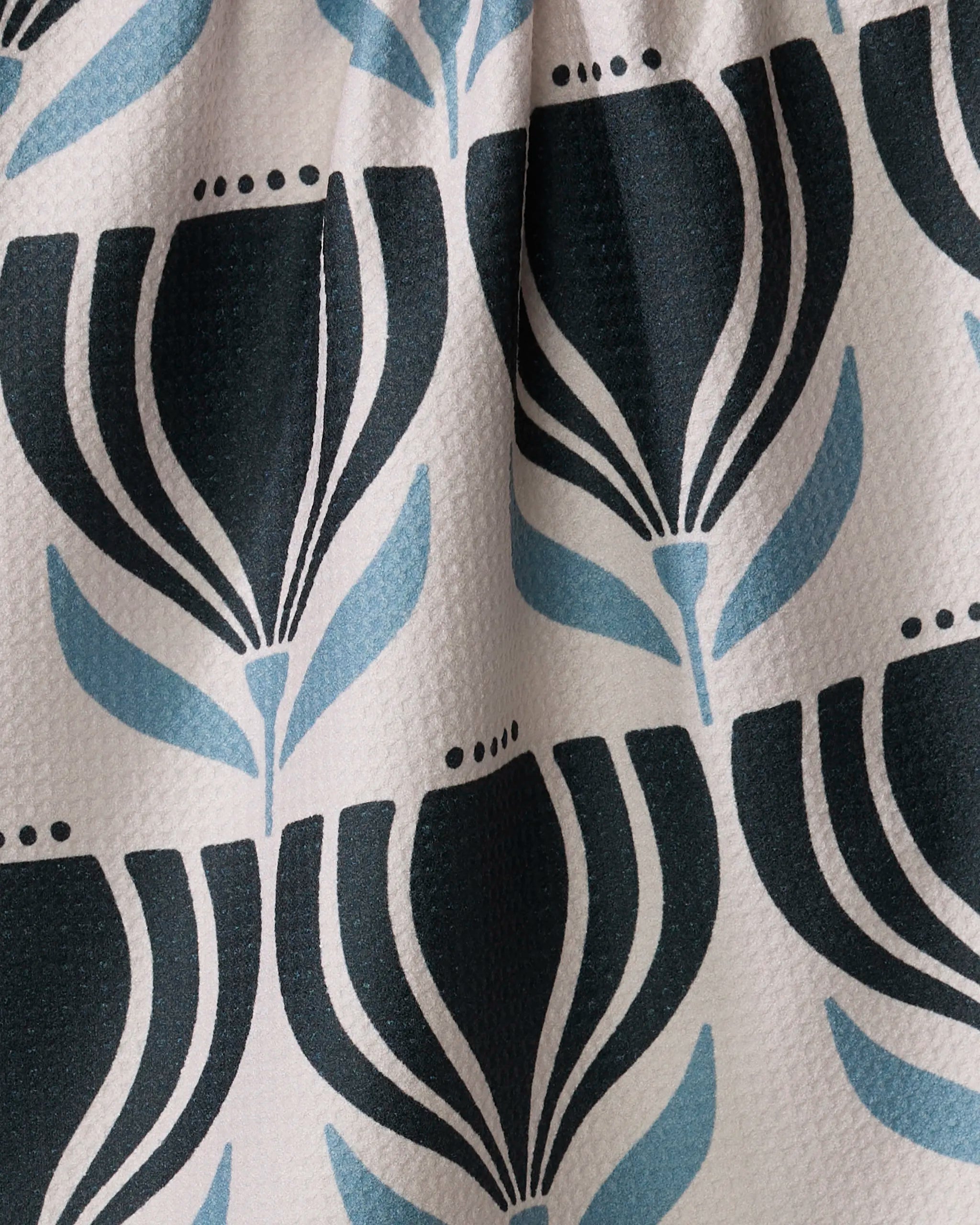 Close-up of the Amy MacCready Florian tea towel, made from recycled materials and featuring a repeating pattern of stylized black and light blue tulip-like flowers and leaves on an off-white textured background.