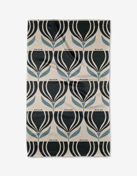 The “Florian” tea towel by Amy MacCready features a repeating geometric pattern of stylized black and blue tulip-like flowers on beige, made from recycled materials for a modern, symmetrical look.