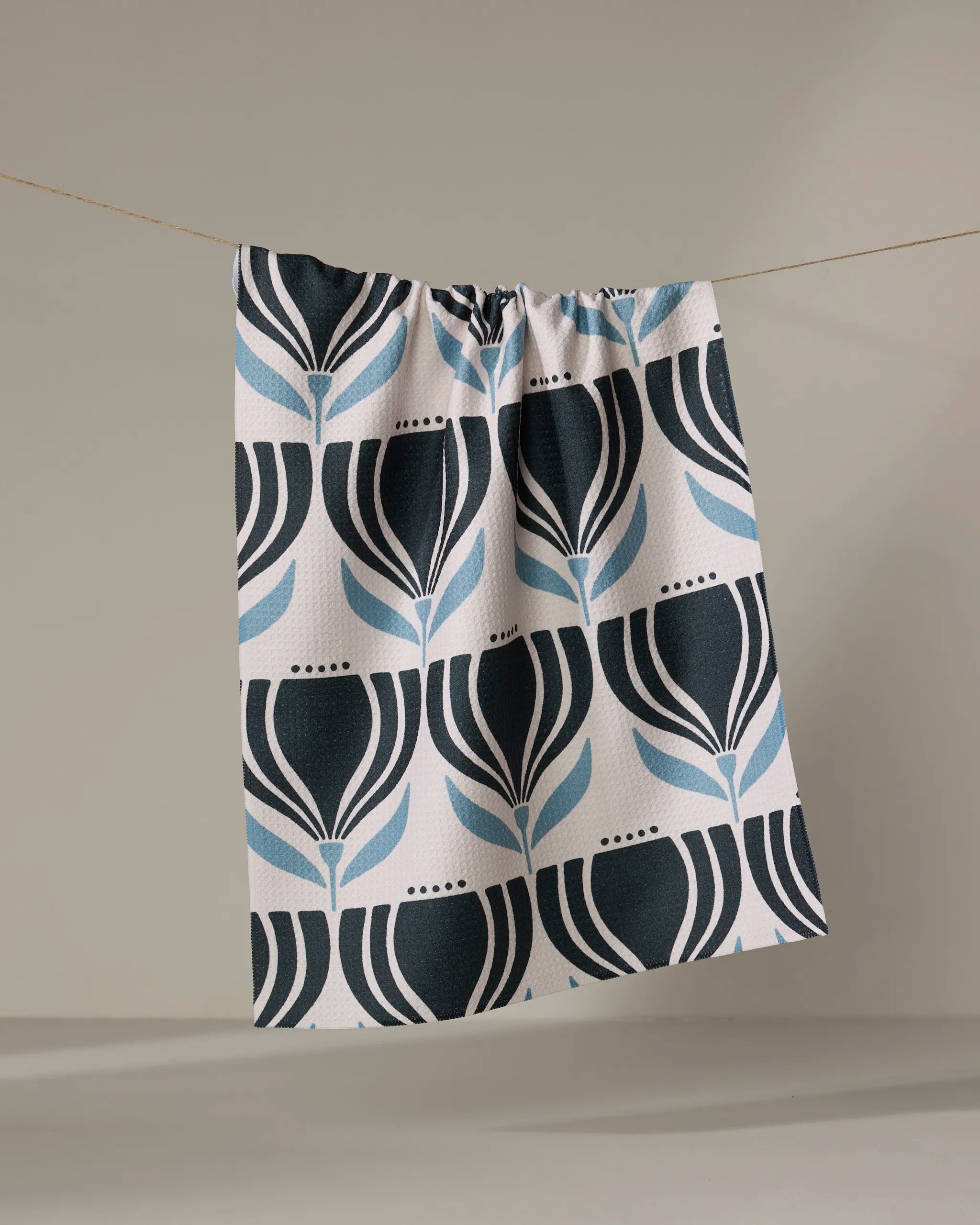 The Florian tea towel by Amy MacCready, made from recycled materials and featuring black and blue floral patterns, hangs on a string against a light gray background.