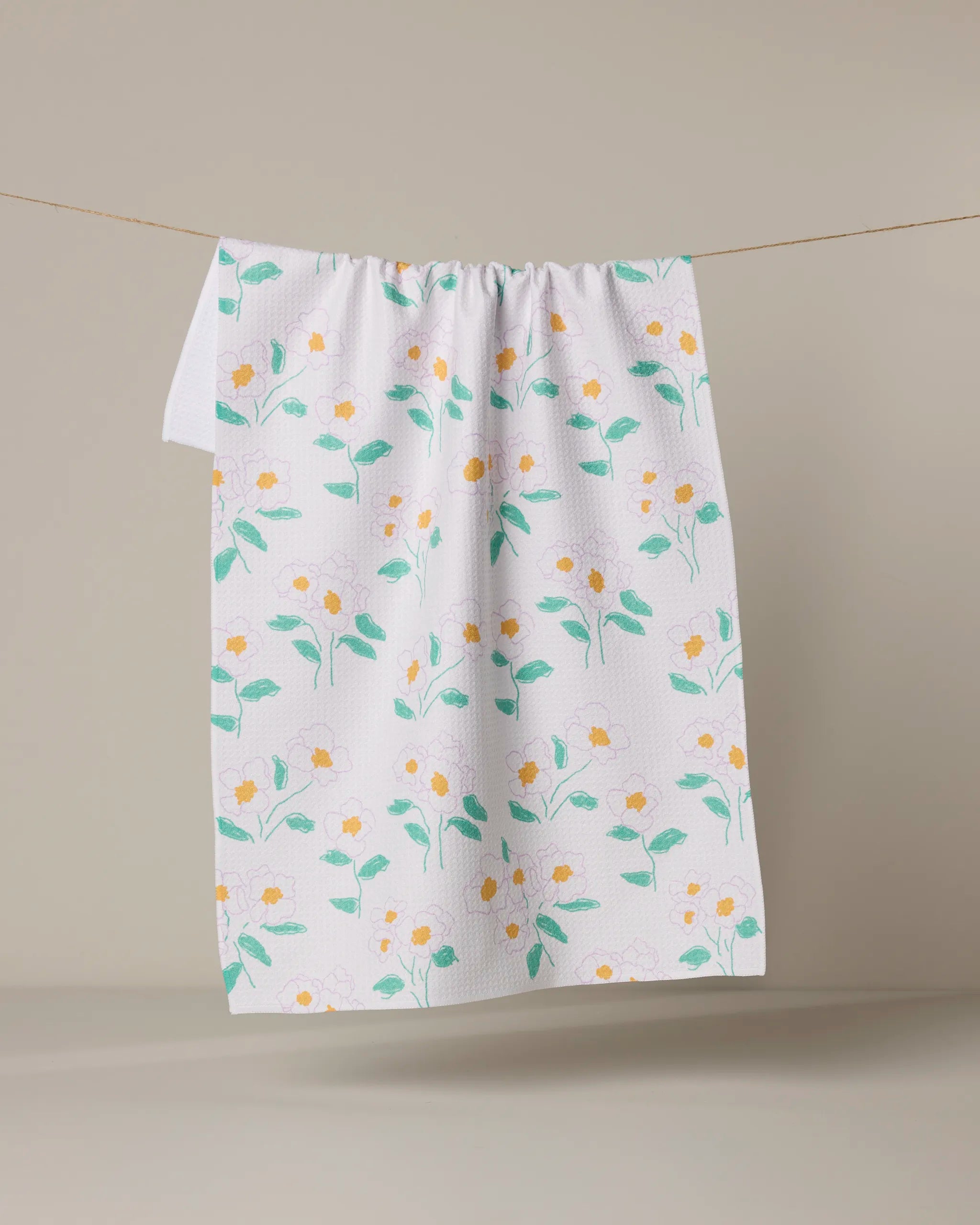The Geometry Florista white tea towel, featuring yellow and white flowers with green leaves, hangs on a line against a beige background.