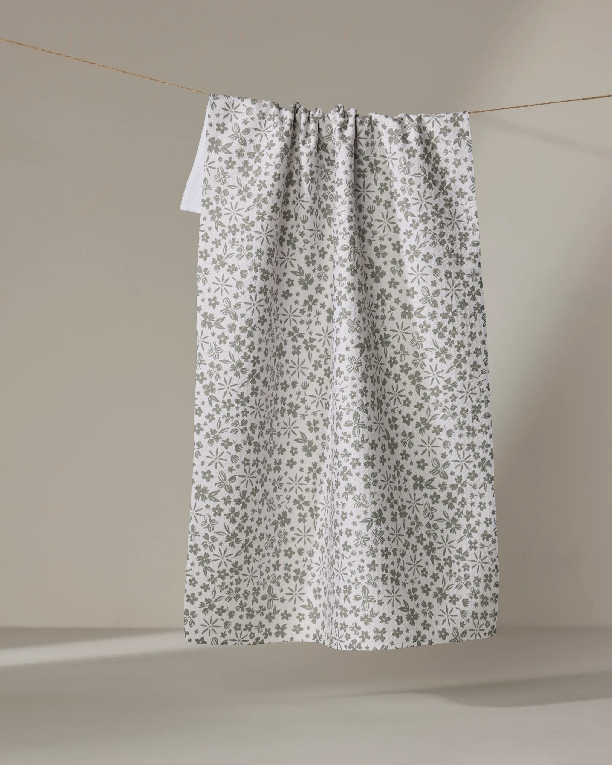 The Flourish Gardens tea towel by Hufton Studio, featuring a subtle grey floral pattern on white, hangs from a thin line against a plain light background.