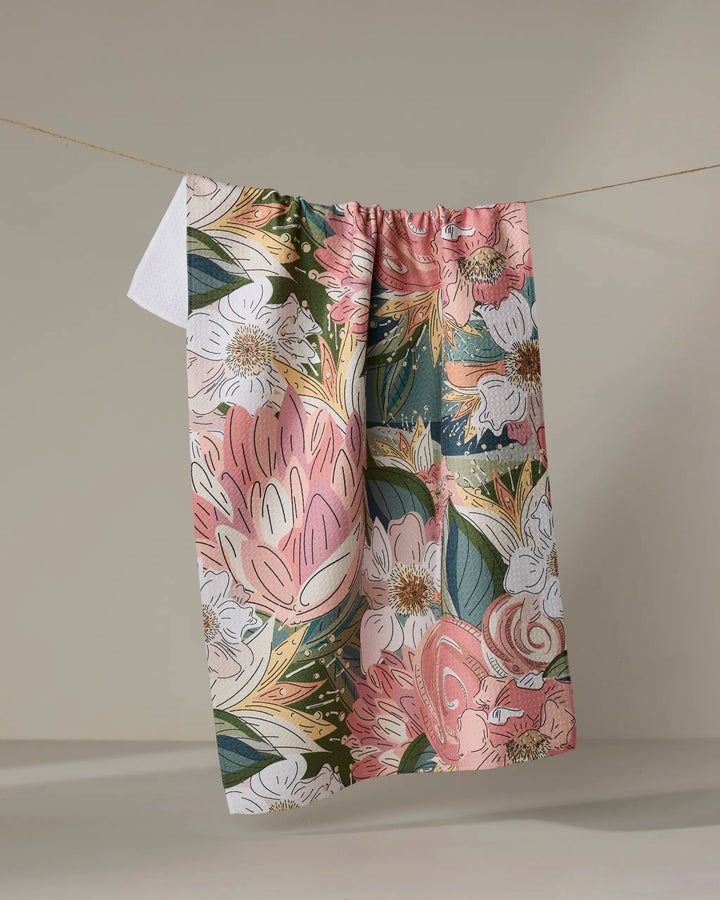 The Flower Explosion tea towel from Second Nature Studio features a vibrant floral print in pink, green, white, and yellow. Made from ultra-absorbent fabric for everyday use, it hangs on a clothesline against a plain beige background.