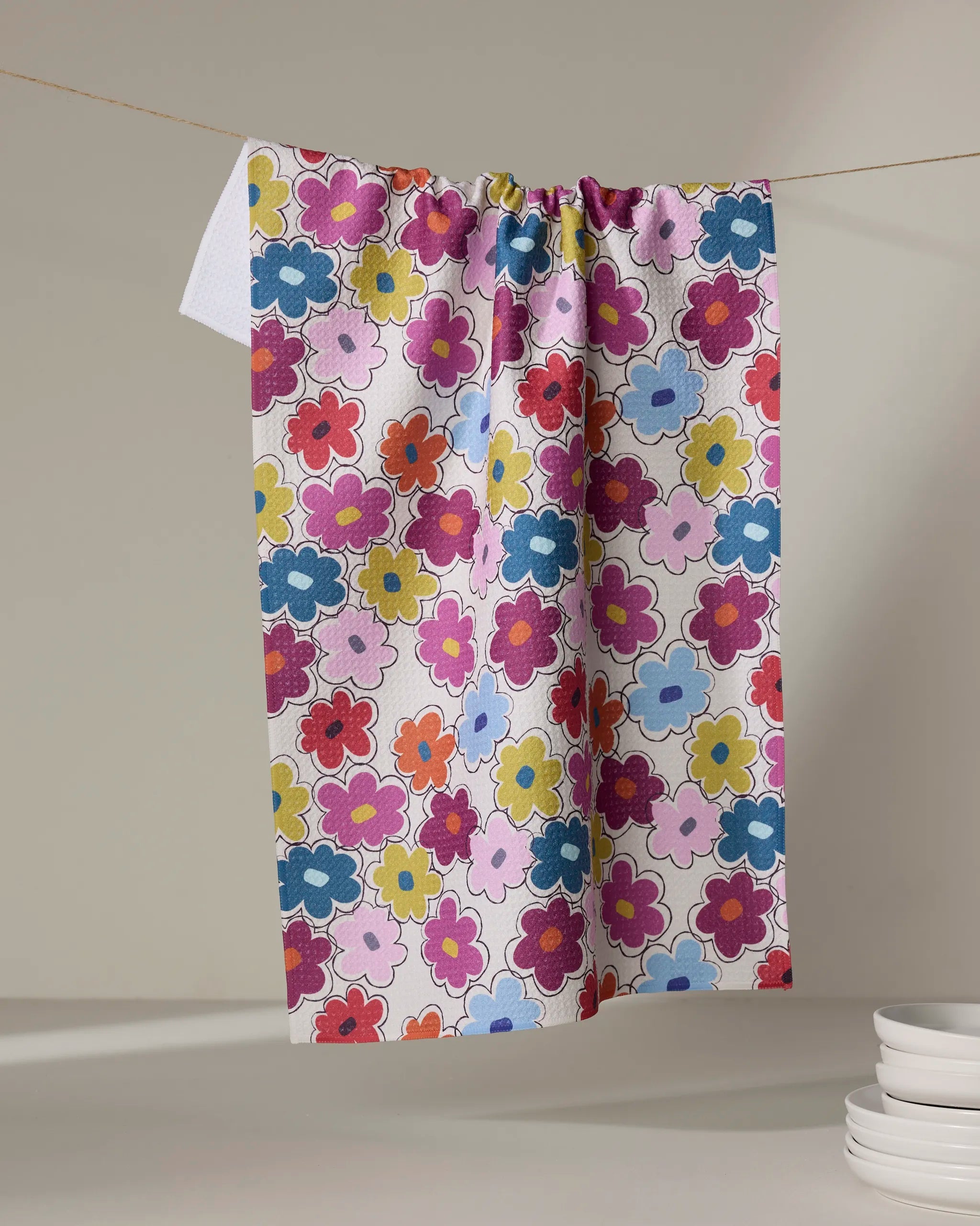 The Lizzie Clark "Flower Fling" tea towel, made from recycled materials, features vibrant red, pink, yellow, blue, and purple flowers. It hangs on a string above a neutral background with stacked white dishes in the lower right corner.