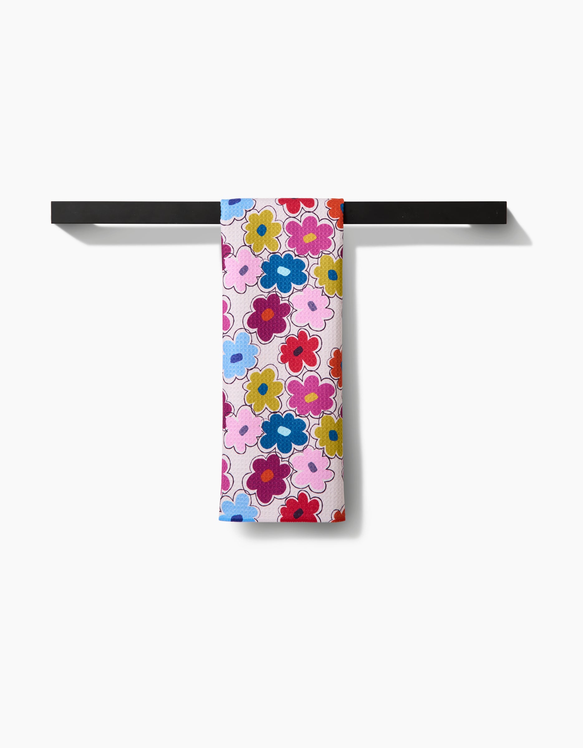 The Lizzie Clark Flower Fling tea towel, featuring a vibrant floral pattern in red, pink, blue, and yellow on white and made from recycled materials, hangs neatly on a black towel bar against a plain white background.