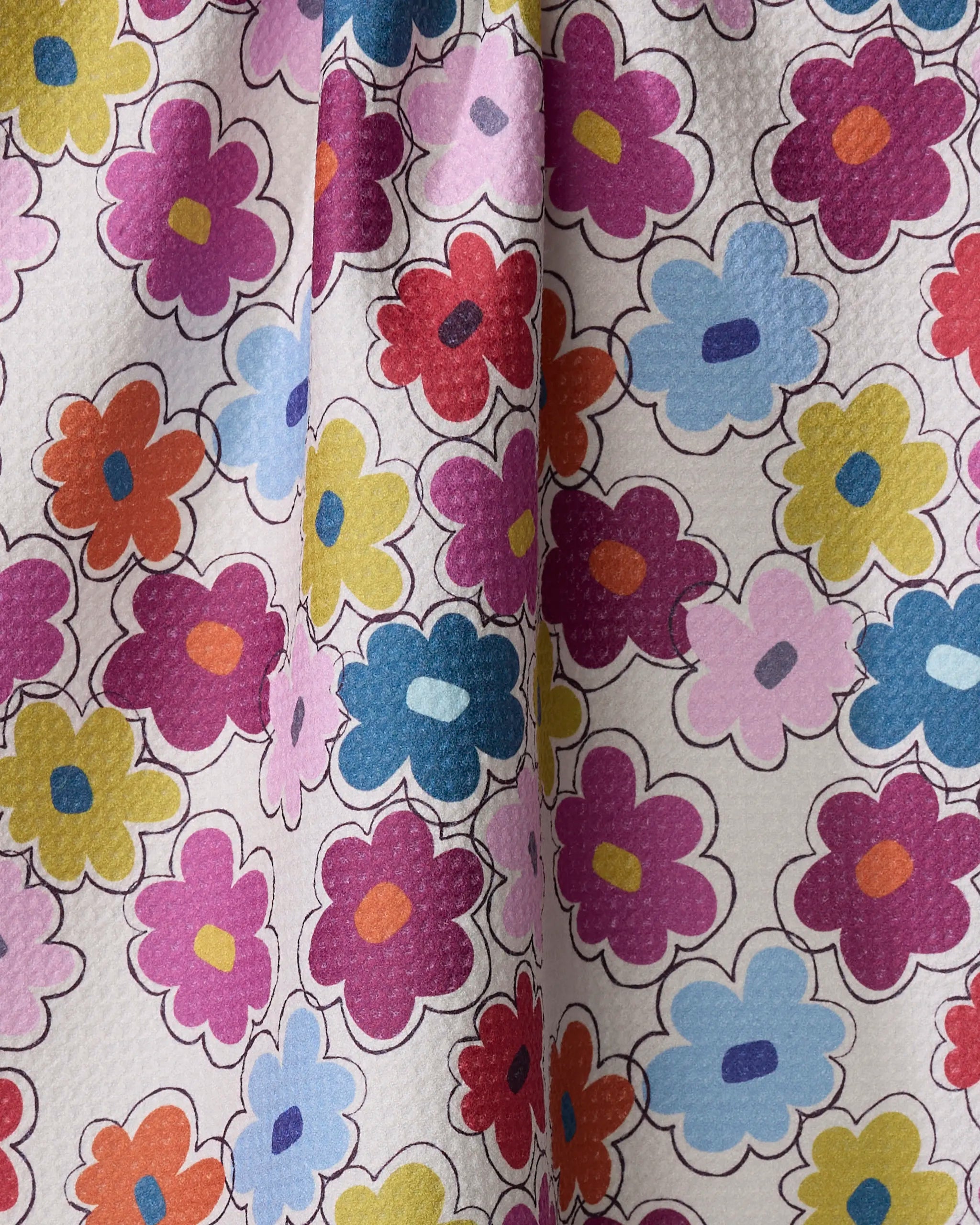 The Flower Fling tea towel by Lizzie Clark features a playful pattern of cartoon-style flowers in pink, red, orange, blue, and yellow with bold outlines on textured white fabric made from recycled materials. Folds and shadows add depth.