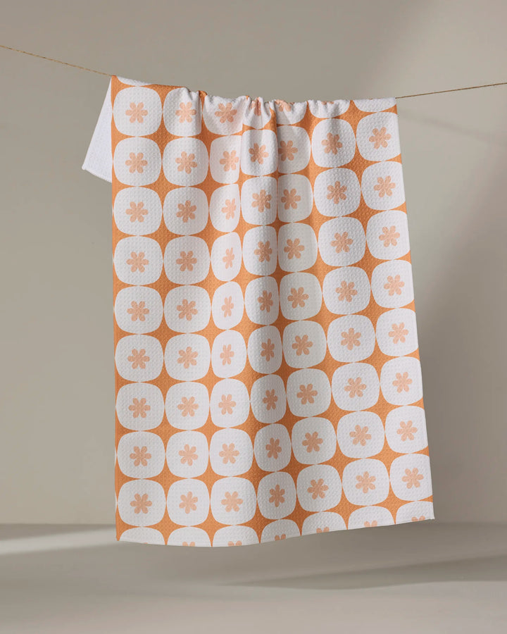 The Flower Power tea towel by Mindfulnice features a peach floral pattern in rounded squares and hangs draped over a clothesline against a neutral background.