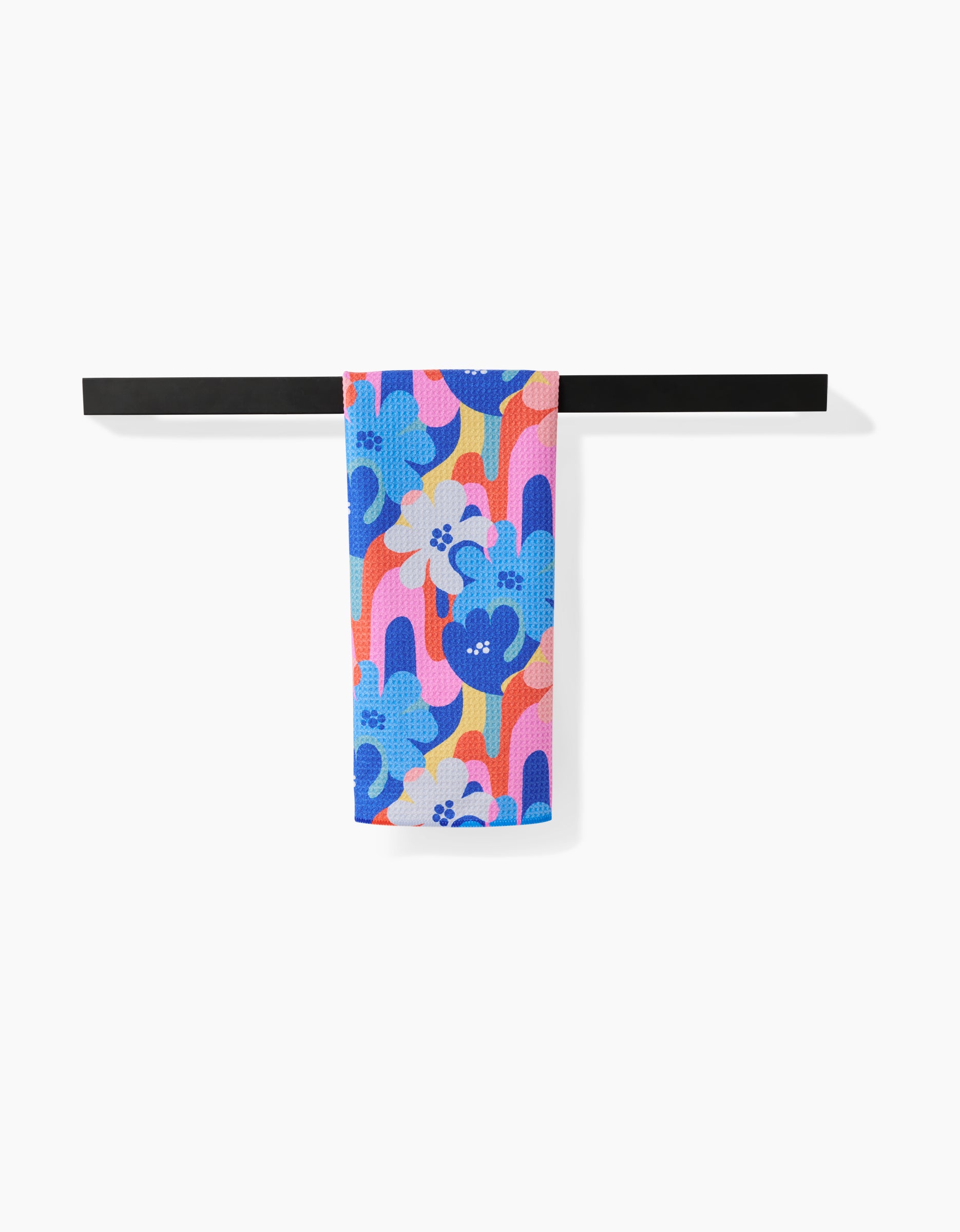 The Masha Volnova Flower Powers tea towel, featuring a striking blue, purple, and orange floral design, is neatly draped over a sleek black towel bar set against a simple white backdrop.