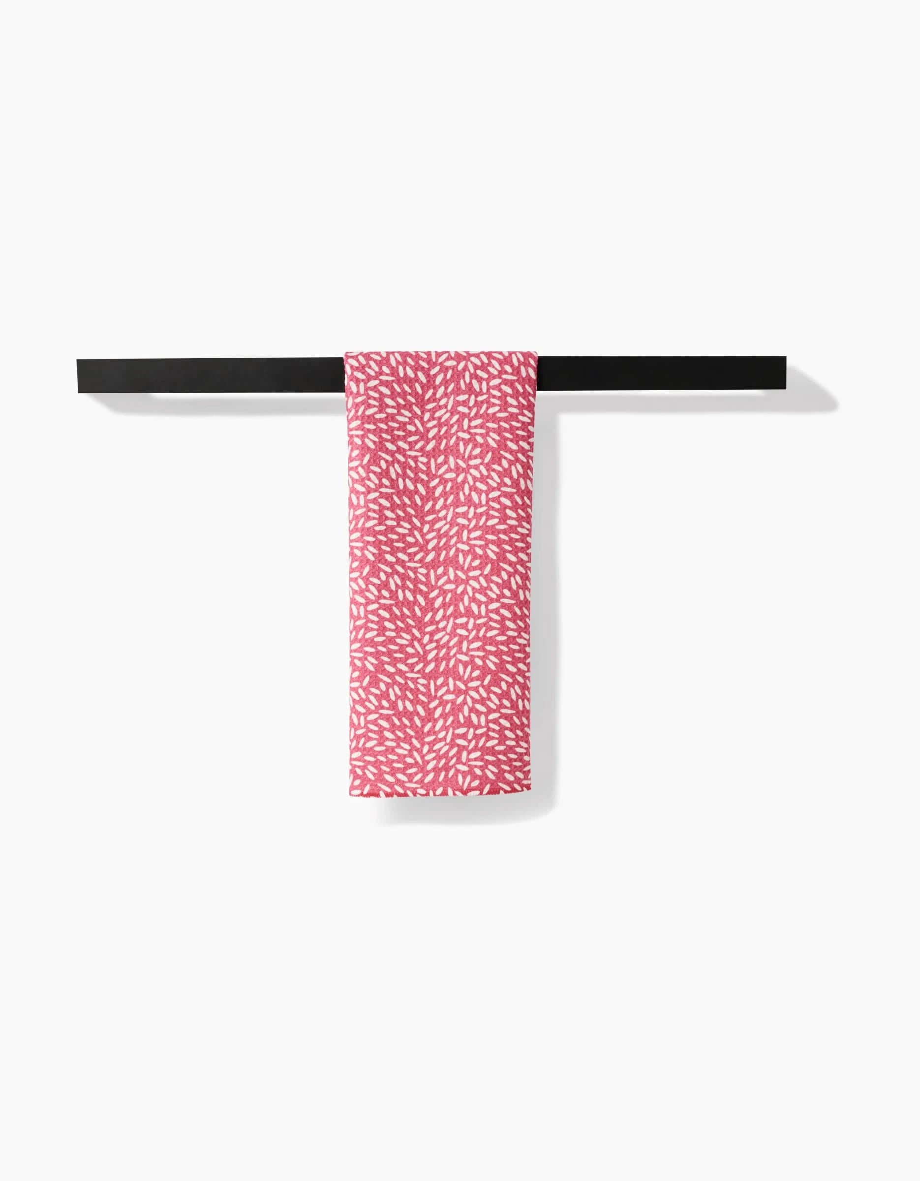 The Flower Seeds tea towel by Amita Nair, featuring a white leaf-like pattern on pink and made from recycled materials, is draped over a simple black towel bar against a plain white background.