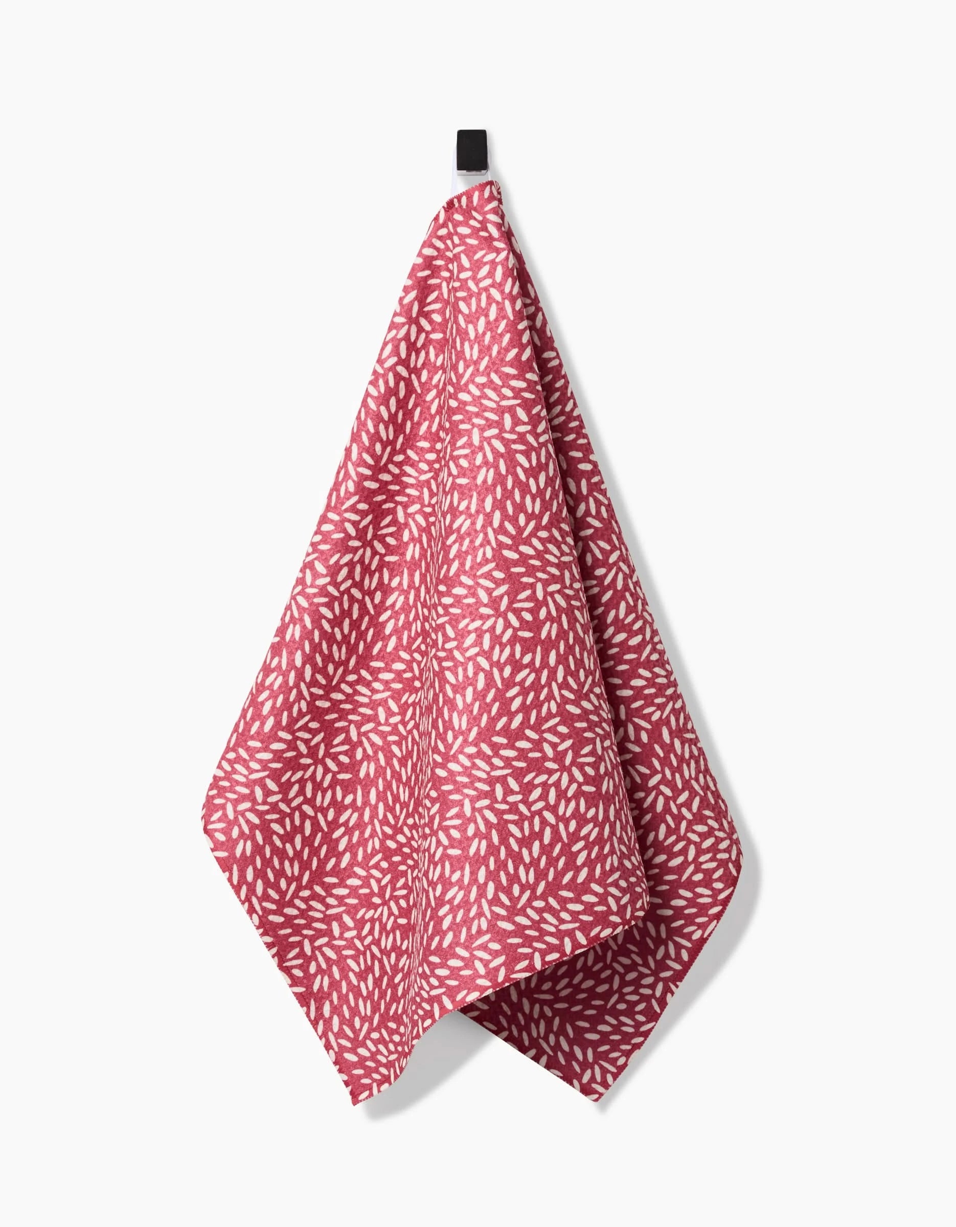 The Flower Seeds tea towel by Amita Nair, featuring a red Geoweave fabric with a white leaf motif, hangs from a black loop against a plain white background.