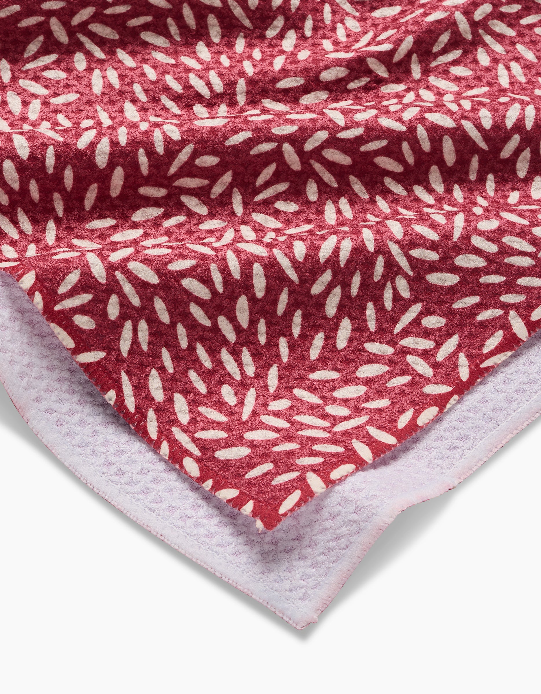A close-up of the Amita Nair Flower Seeds tea towel, red with a white leaf pattern and a light purple textured underside, partially folded to display both sides.