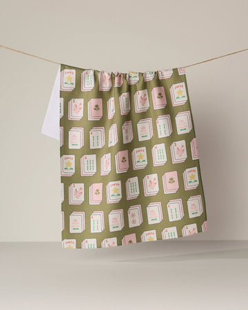 A green and pink patterned tea towel from Geometry, called "Flower Tiles," featuring a Mahjong-inspired design, hangs on a string against a neutral background, bringing an eco-friendly accent to your kitchen decor.