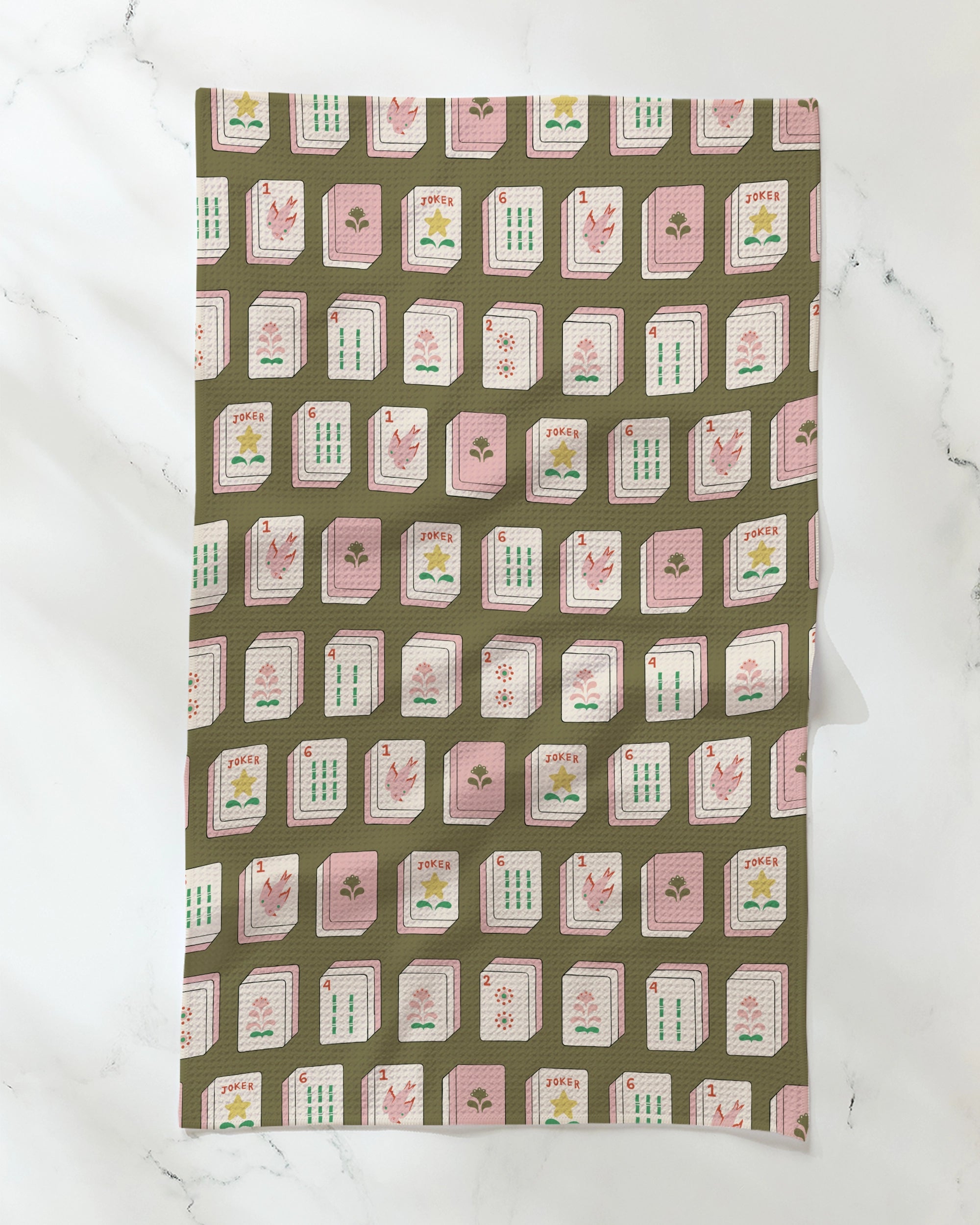 A brown, eco-friendly tea towel from Geometry, called “Flower Tiles,” featuring a repeating pattern of Mahjong tiles in pink, white, and green, displayed on a white marble surface.